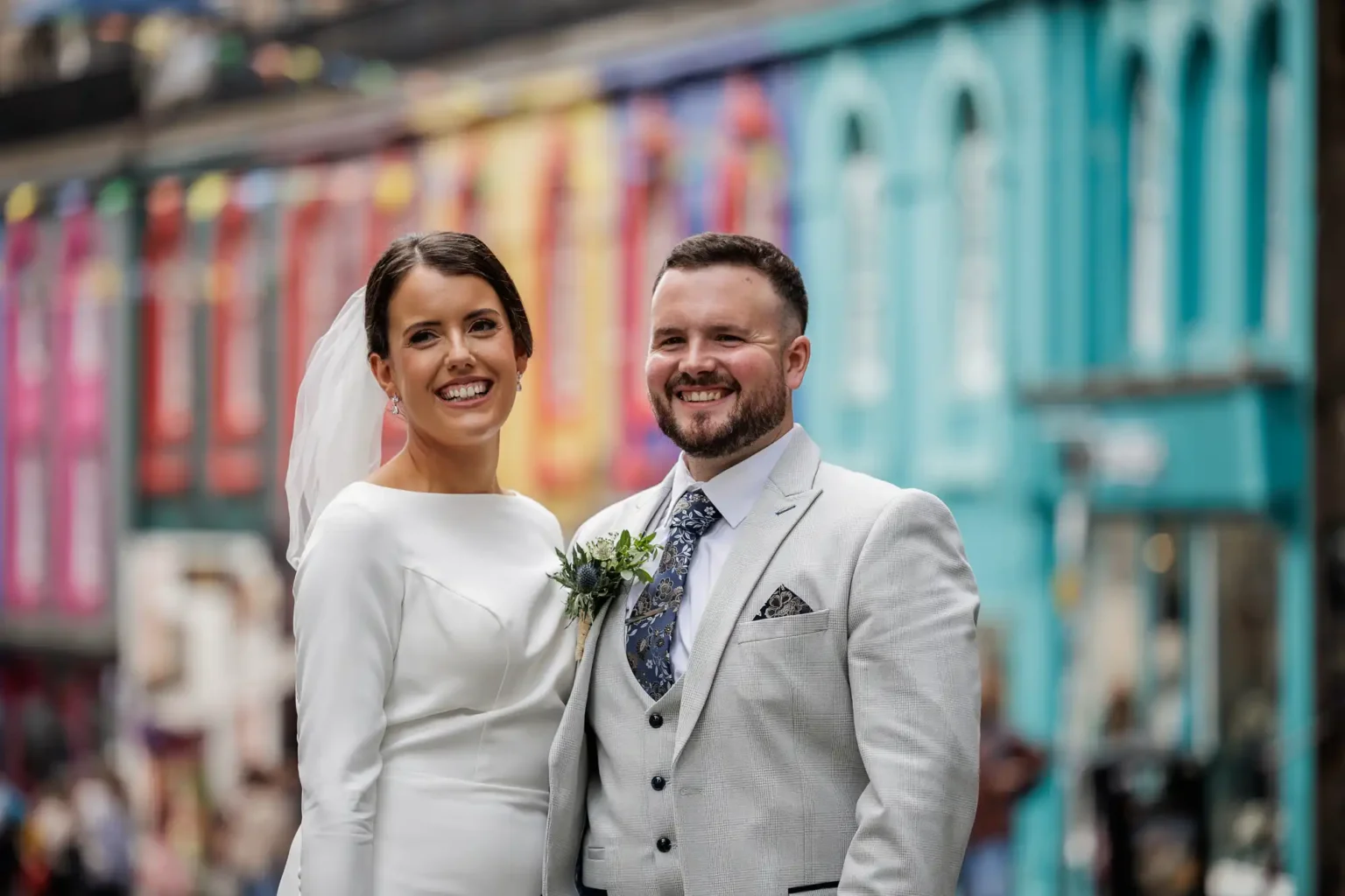 Edinburgh City Chambers micro wedding photo of a smiling bride in a white dress and veil stands next to a groom in a light grey suit and tie. They pose outdoors on a street with colourful, blurred buildings in the background, creating a festive, joyful atmosphere.