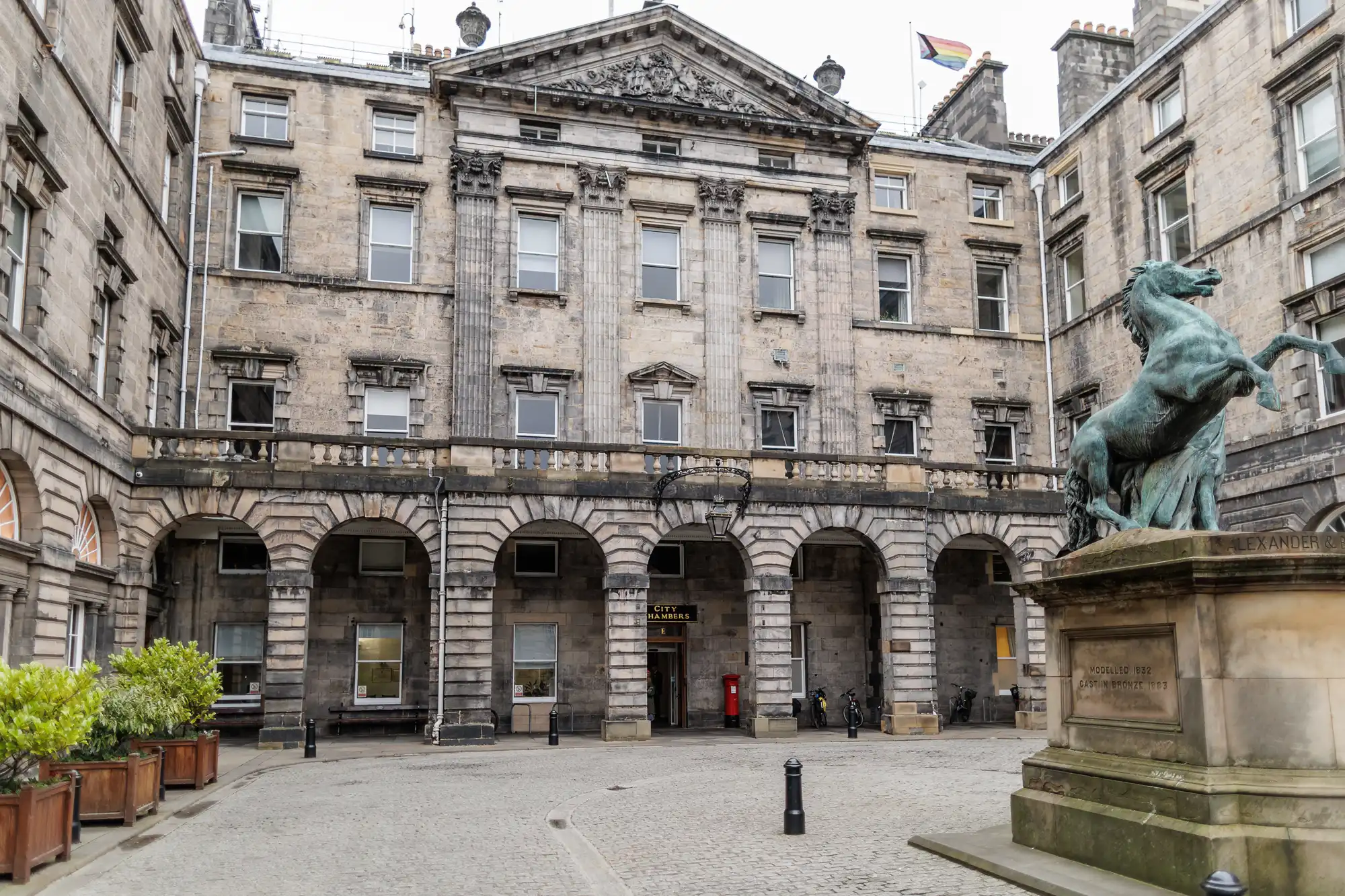A historic stone building with arched entrances and classical columns stands in a cobbled courtyard. A bronze horse statue on a plinth is visible to the right. Several windows line the façade, and a rainbow flag is flown from the roof.