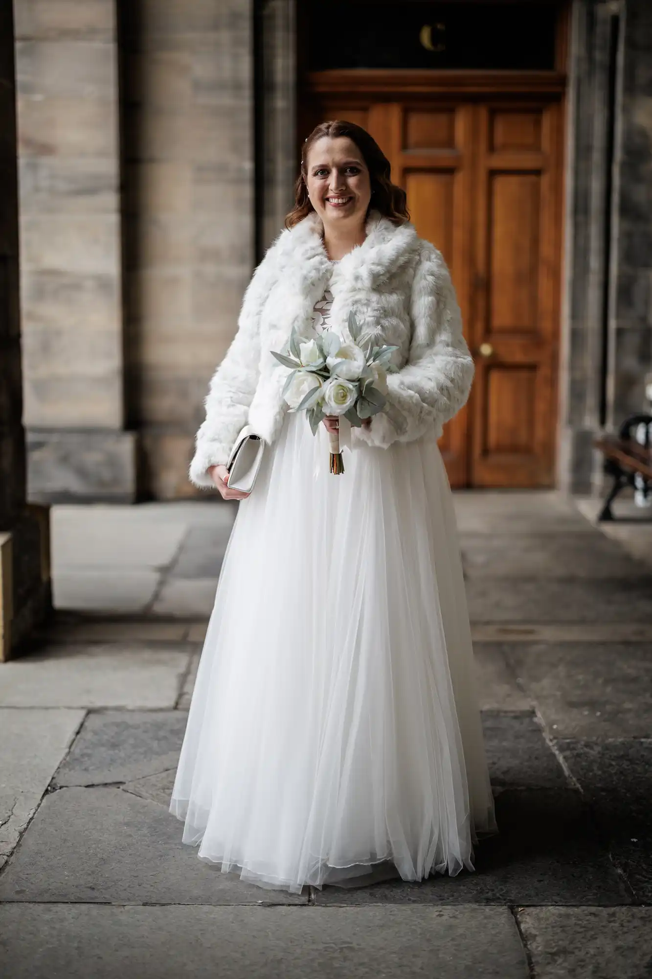 A bride stands outdoors on stone pavement, smiling. She wears a white wedding dress with a tulle skirt and a white faux fur jacket. She holds a bouquet of white flowers and a clutch. Behind her is a wooden door with stone walls on each side.