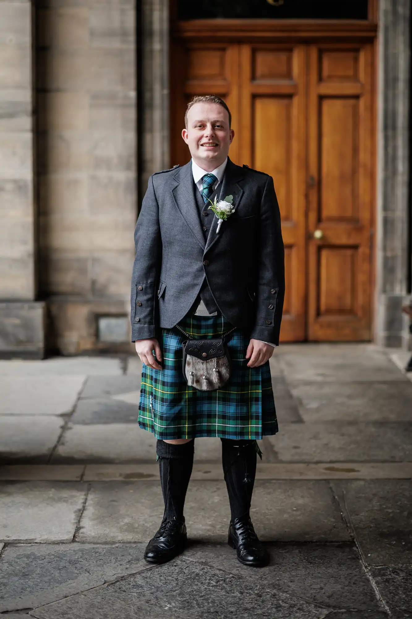 A smiling man stands outside in front of wooden double doors, wearing a traditional Scottish outfit: a grey jacket, green and blue tartan kilt, sporran, knee-high socks, and black shoes. He has short hair and a buttonhole pinned to his lapel.