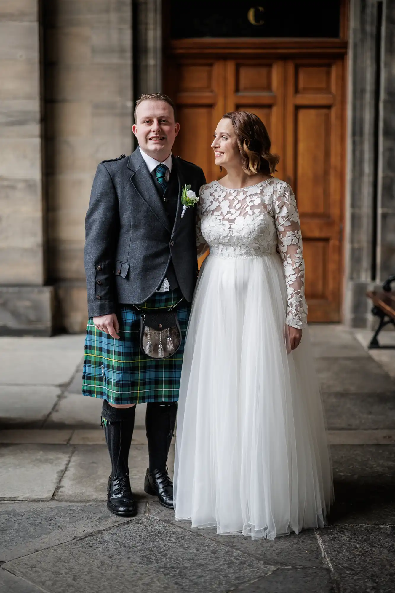 A smiling bride in a white lace-sleeved gown stands beside a groom in a grey jacket, green tartan kilt, sporran, and tie. They pose happily in front of a wooden door set in a stone building.