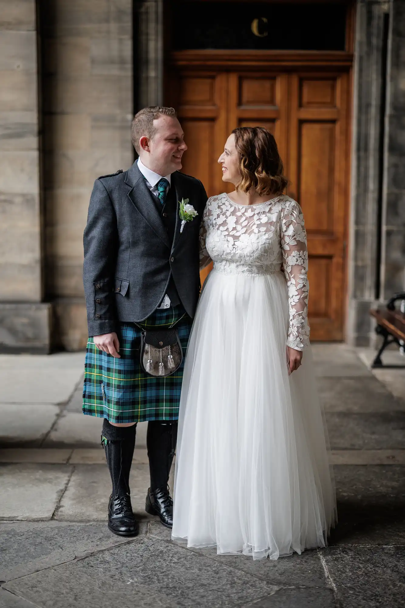 A bride in a white lace and tulle gown stands smiling beside a groom wearing a dark jacket, tartan kilt, sporran, long socks, and shoes. They face each other, holding hands, with a wooden door and stone walls in the background.