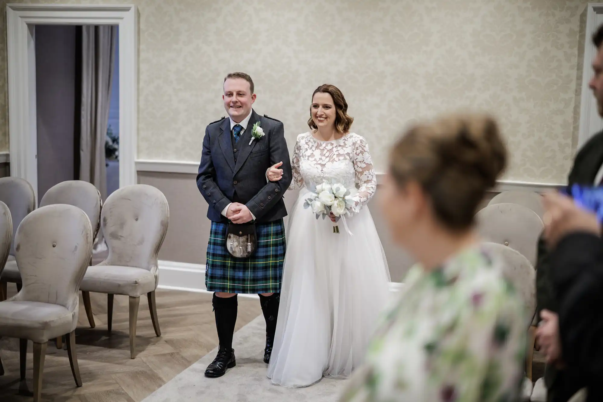 A smiling bride in a long white dress with lace sleeves holds a bouquet and walks arm-in-arm with a man in a dark jacket and blue tartan kilt down an aisle in an elegant room with beige chairs and light wallpaper. Guests are blurred in the foreground.