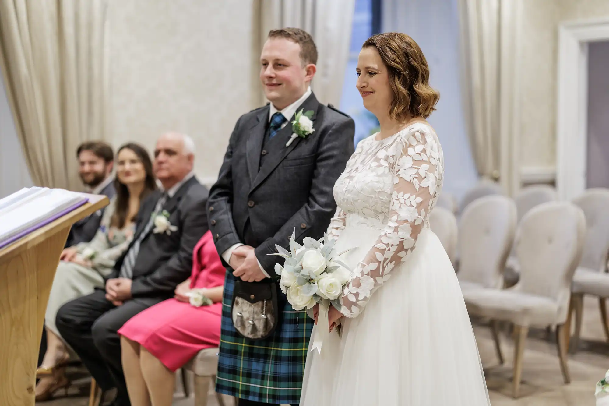 A bride in a white dress with lace sleeves and a groom in a tartan kilt stand together, holding flowers, during a wedding ceremony. Four seated guests watch them in a softly lit room with cream walls and curtains.