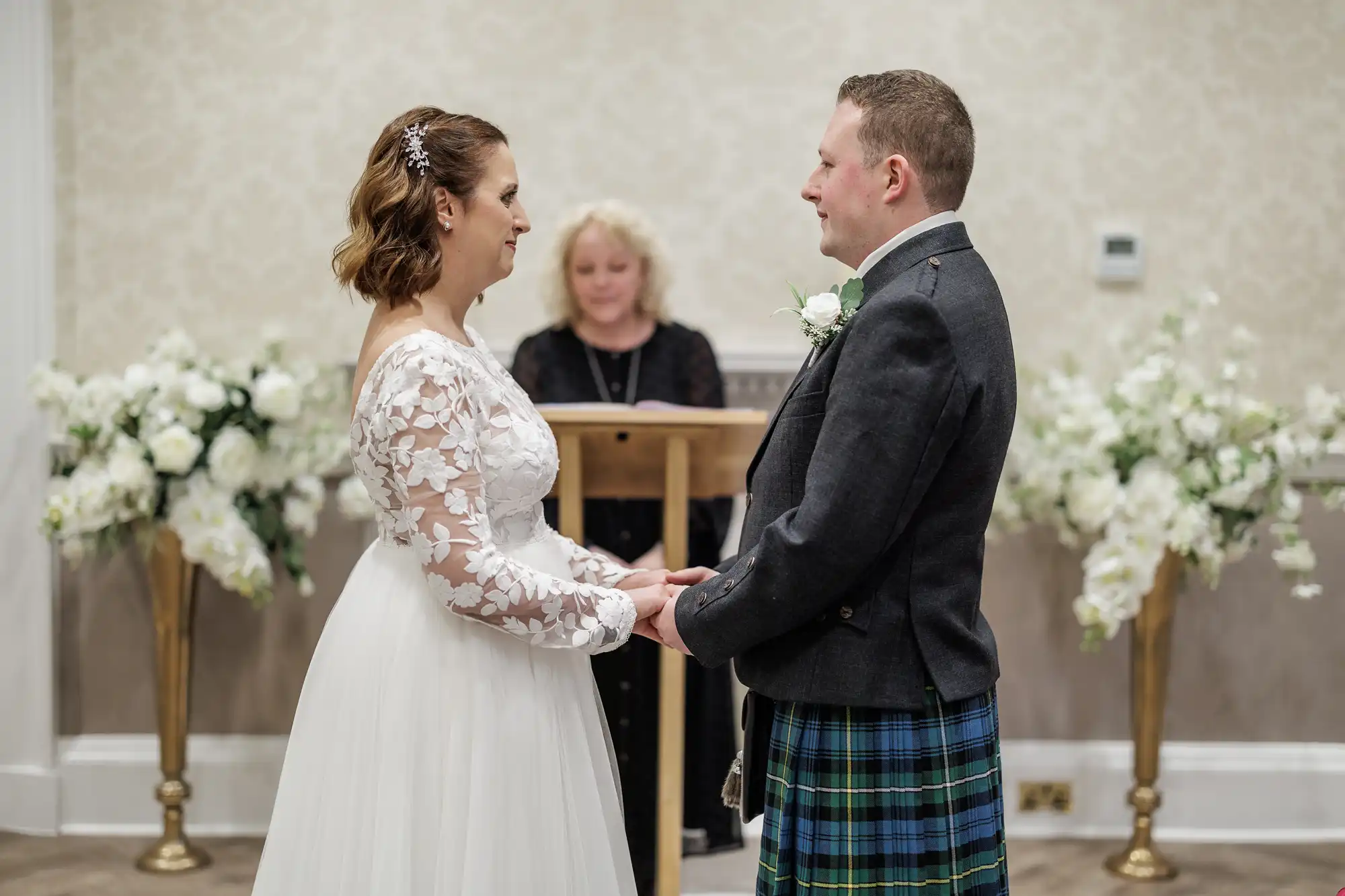 A bride in a long-sleeved lace gown and a groom in a dark jacket with a tartan kilt hold hands facing each other. They smile during a wedding ceremony, with a woman officiating behind them and large white floral arrangements in the background.