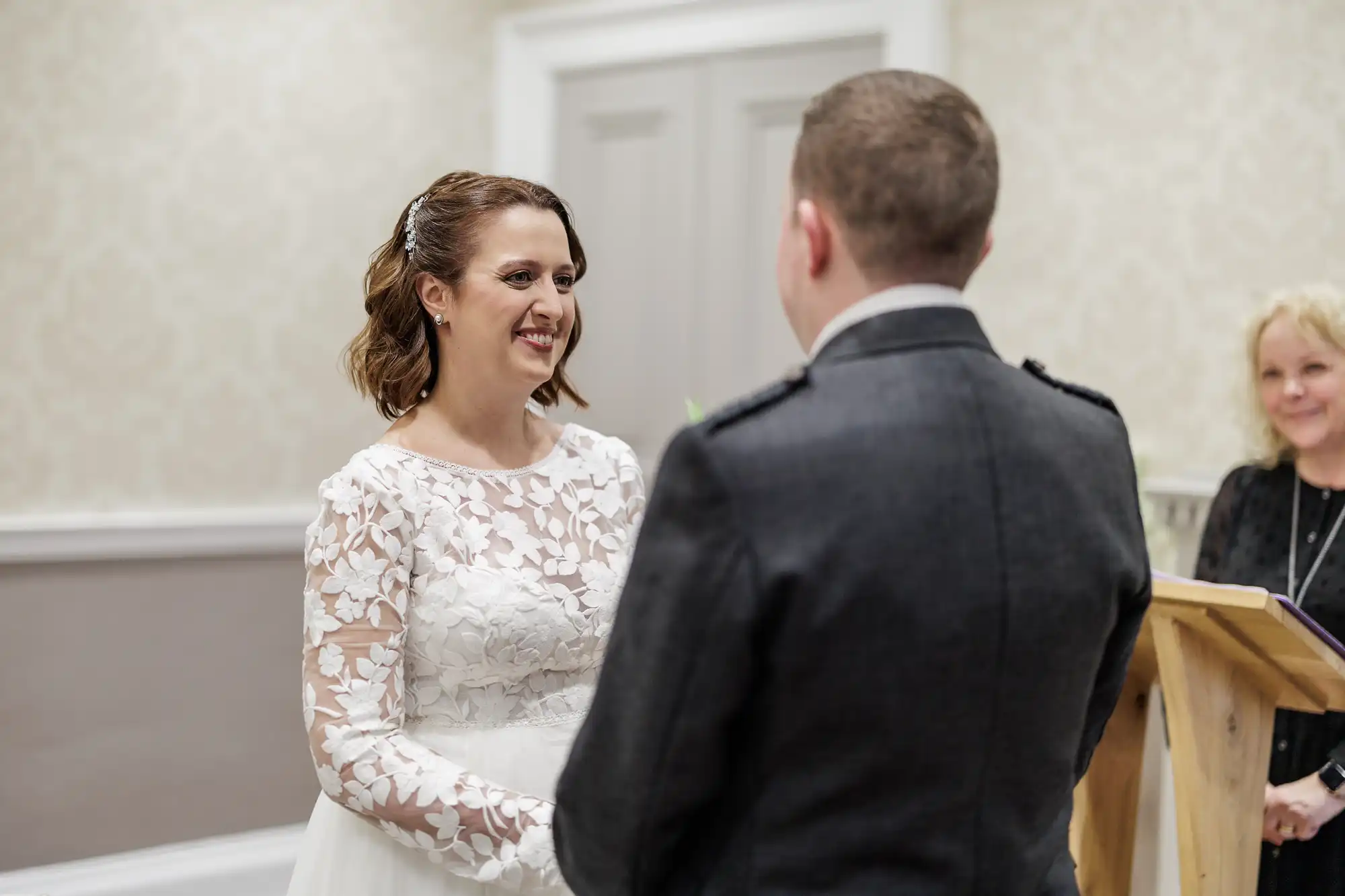 A bride in a white lace dress smiles as she holds hands with a groom in a dark suit. They stand facing each other indoors, with a woman officiant in the background by a wooden lectern. The setting is softly lit with neutral-toned walls.