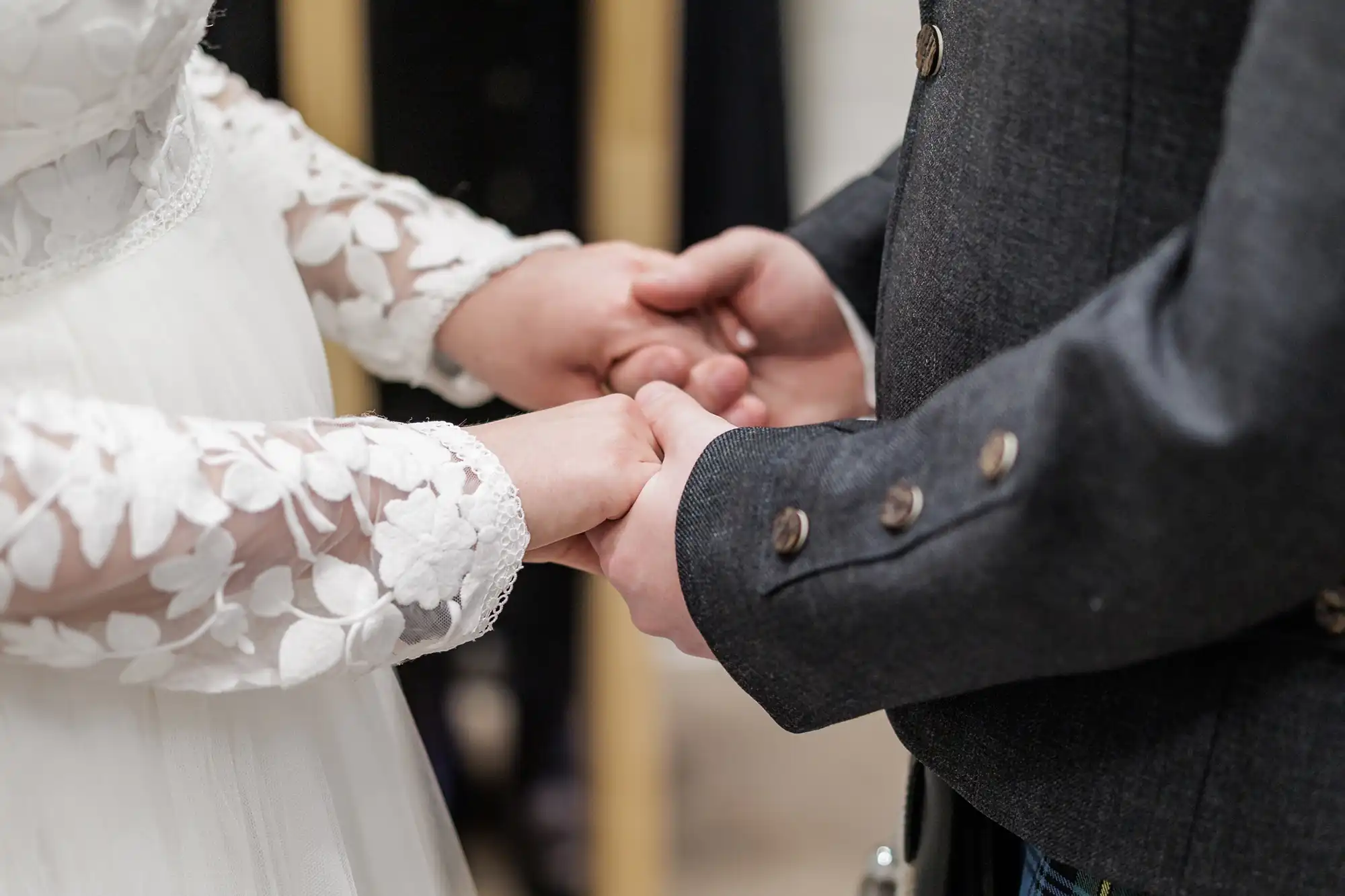 A close-up of a bride and groom holding hands. The bride wears a white dress with floral lace sleeves, and the groom wears a dark suit with silver buttons. Their hands are gently clasped, focussing on their connection. The background is softly blurred.