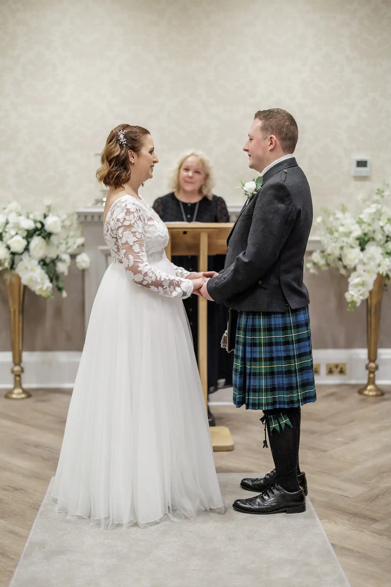 A bride in a white gown with lace sleeves and a groom in a dark jacket and blue tartan kilt stand holding hands, facing each other during their wedding ceremony. An officiant stands at a lectern behind them, with white floral arrangements on either side.