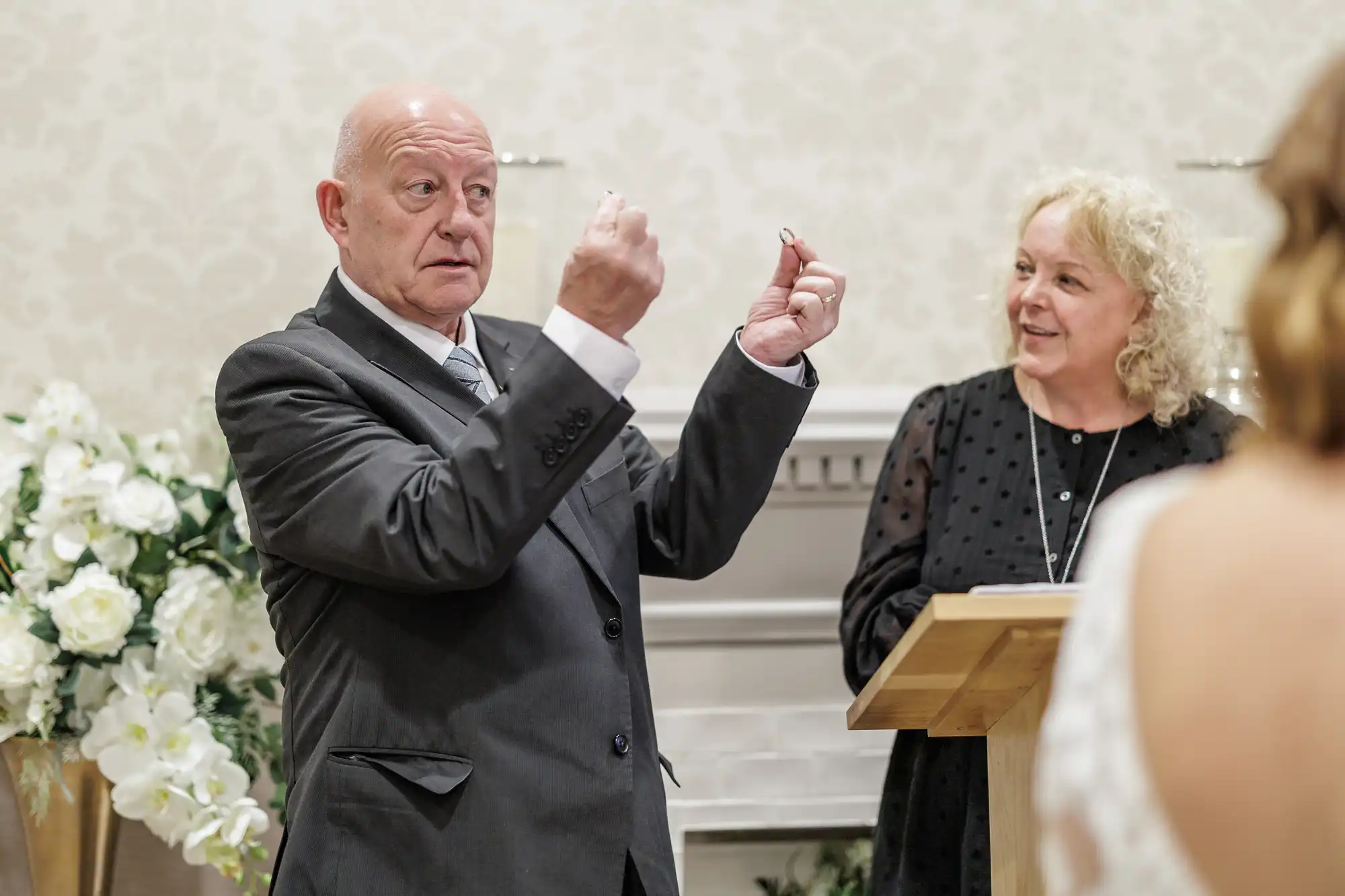 A bald man in a suit holds two rings up while speaking, standing next to a smiling woman in a black dress at a lectern. White flowers and a decorative fireplace are in the background. The back of a bride's dress is visible in the foreground.