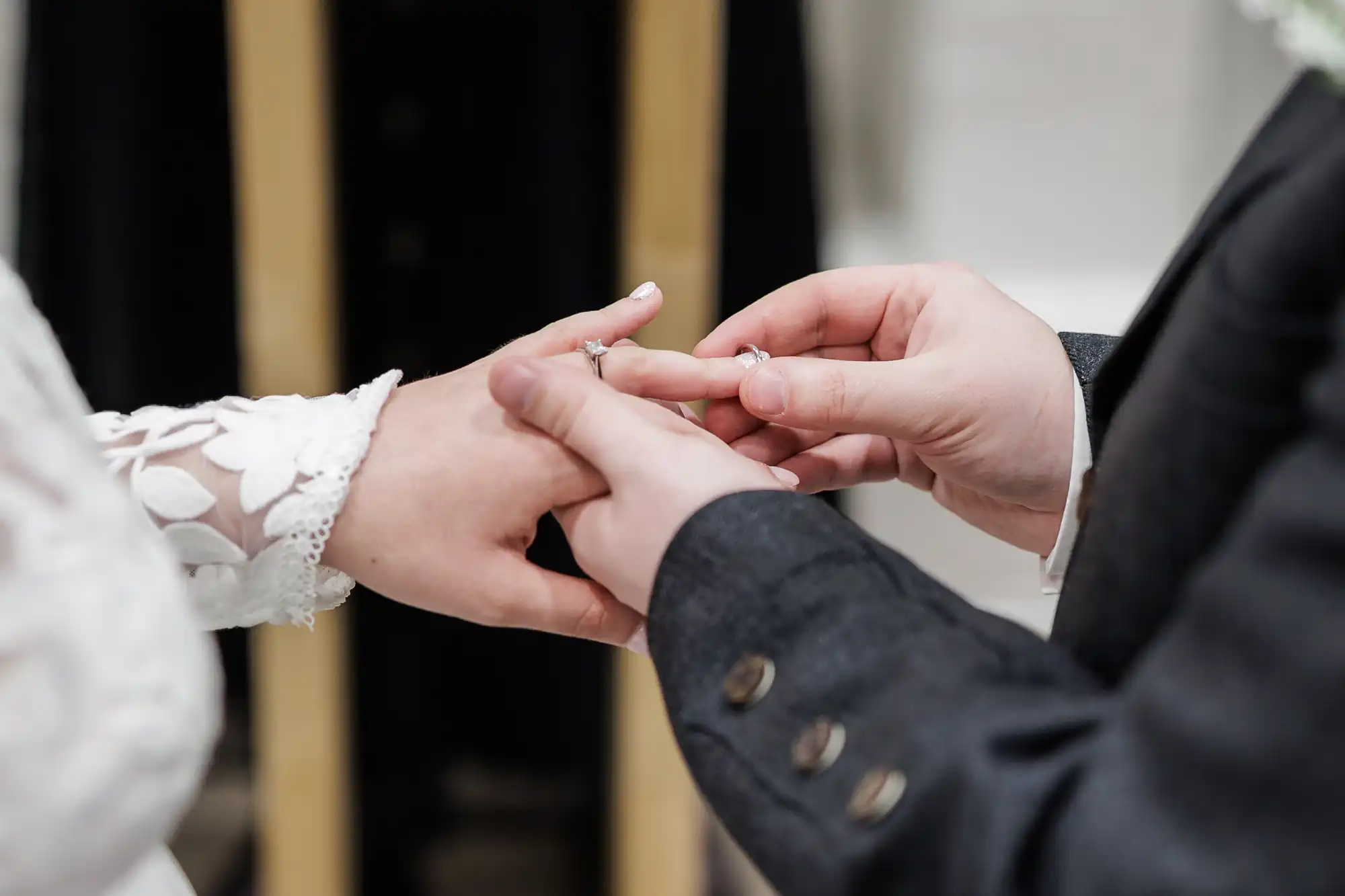 Close-up of one person placing a ring on another's finger during a wedding ceremony. The person receiving the ring wears a white, lace-sleeved dress, and the other wears a dark suit jacket with visible buttons. The background is out of focus.