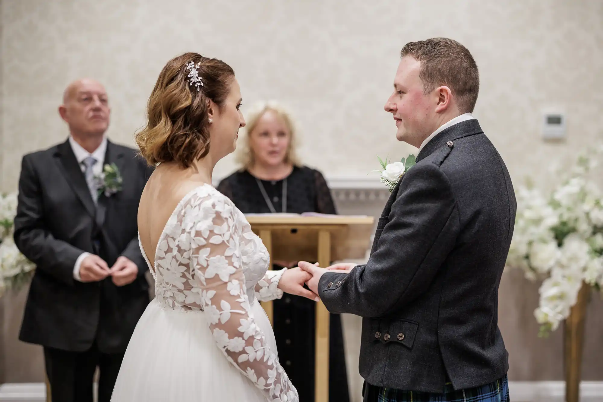 A bride in a white lace dress and a groom in a dark suit hold hands facing each other during a wedding ceremony. An officiant stands behind a lectern, and another man stands to the left. Floral arrangements decorate the background.