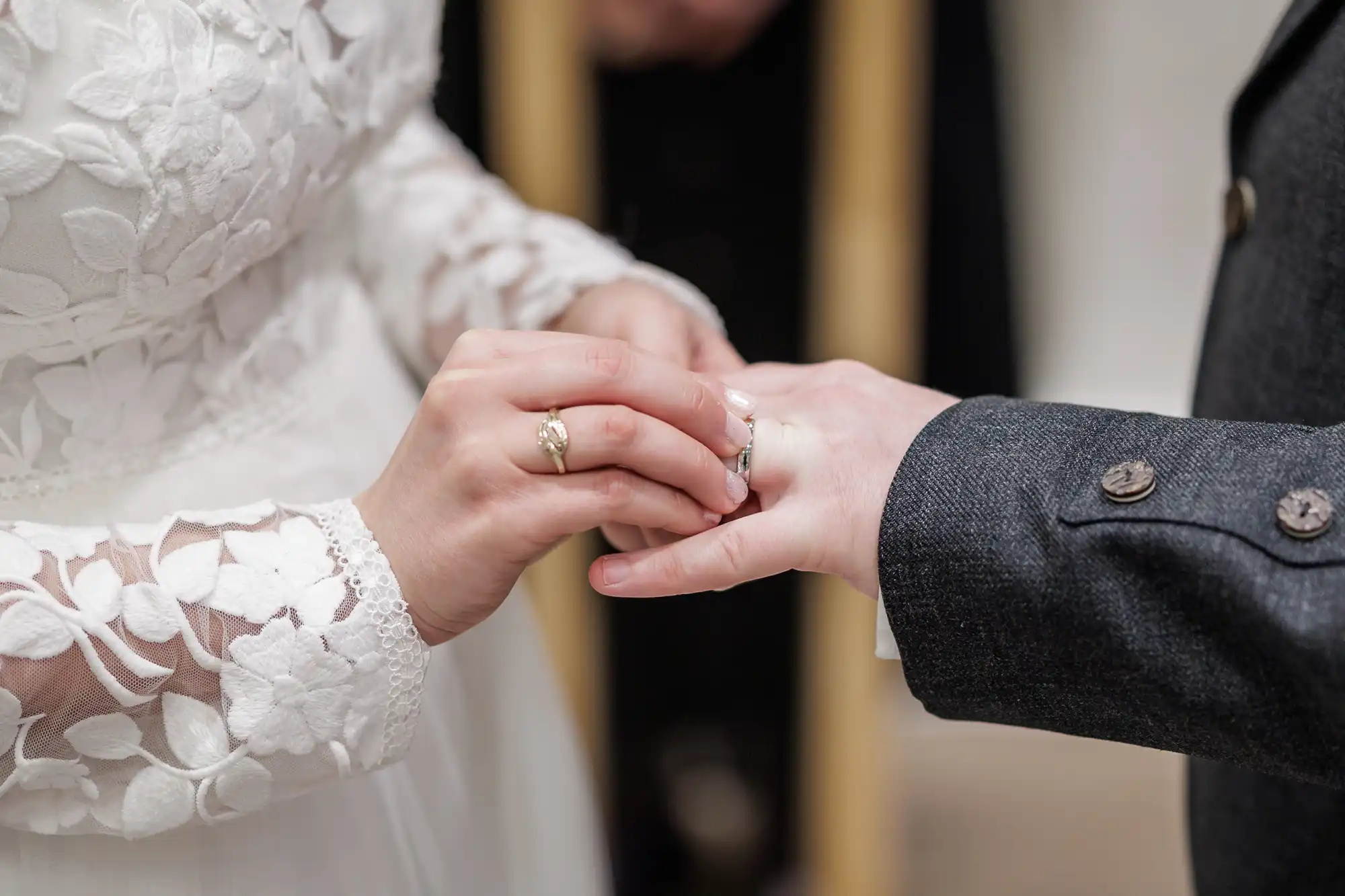 A close-up of a bride in a white, floral lace dress placing a ring on the groom’s finger. The groom is wearing a dark suit with visible buttons. The focus is on their hands during the wedding ceremony. The background is softly blurred.
