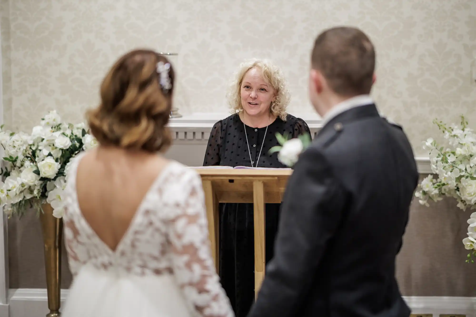 A woman stands at a wooden lectern, smiling as she officiates a wedding ceremony. In the foreground, a bride in a white lace dress and a groom in a dark suit hold hands, seen from behind. White floral arrangements decorate the elegant room.