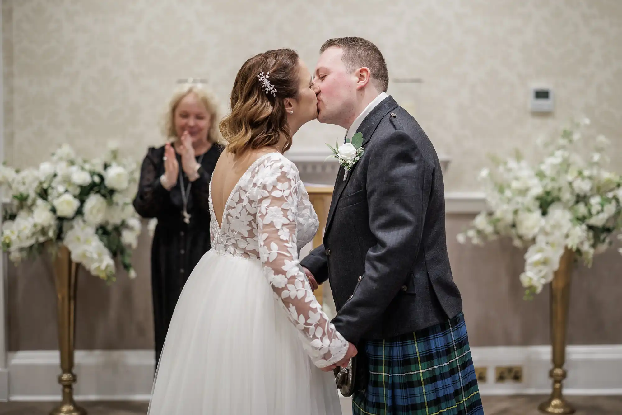 A bride in a white lace-sleeved dress and a groom in a kilt and jacket kiss at their wedding ceremony, holding hands. White floral arrangements flank them, and a smiling woman in a black dress applauds in the background.
