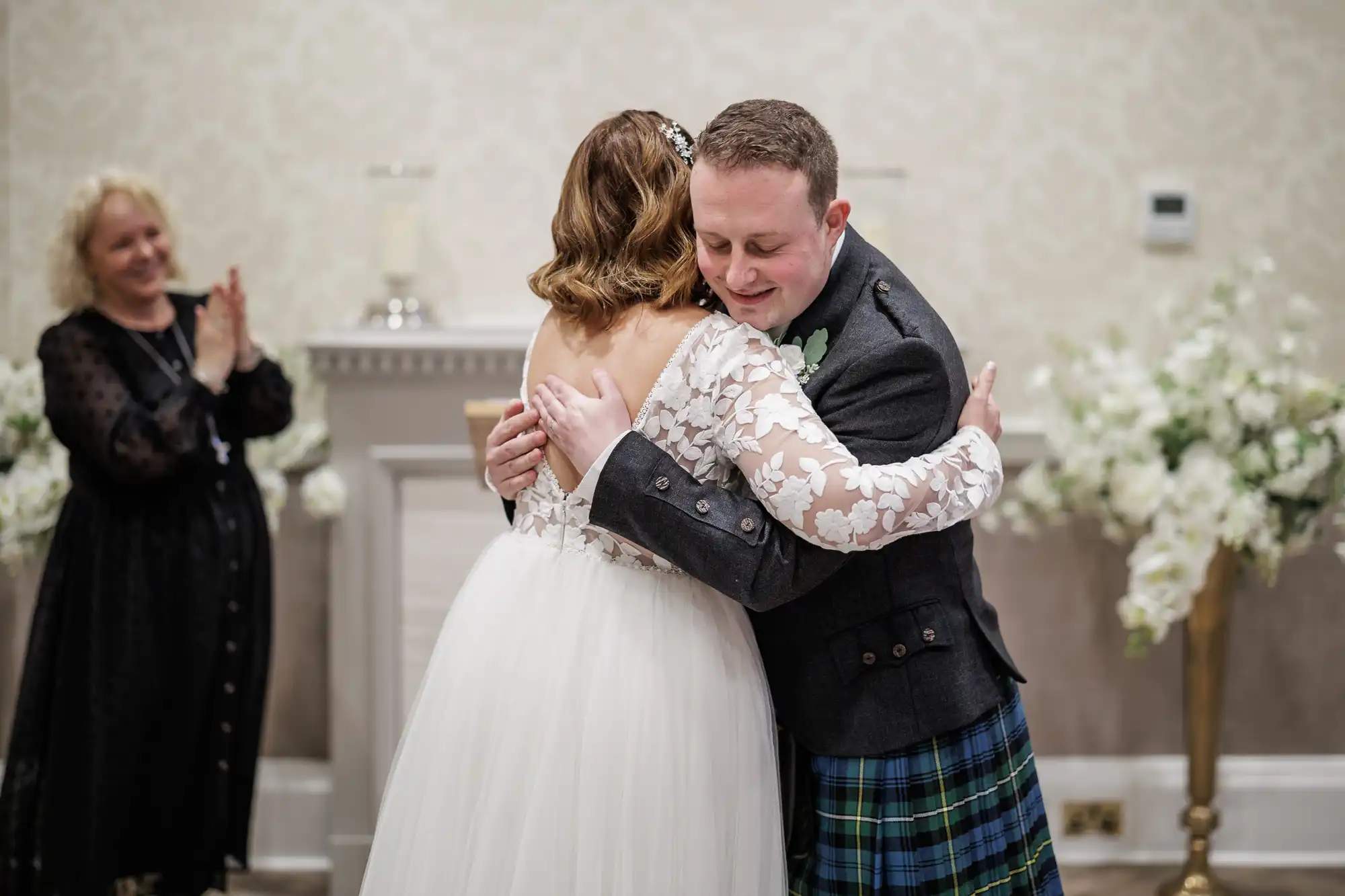 A bride in a white lace dress hugs a groom in a dark jacket and tartan kilt. Both look happy and emotional. An older woman in a black dress claps in the background. White flowers and a decorated fireplace complete the wedding scene.