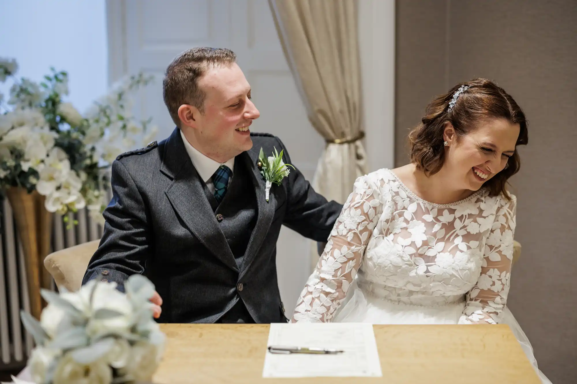 A smiling bride and groom sit together at a table, holding hands. The bride wears a white dress with floral lace, and the groom wears a dark suit with a buttonhole. A pen and paper lie before them, with floral decorations in the background.