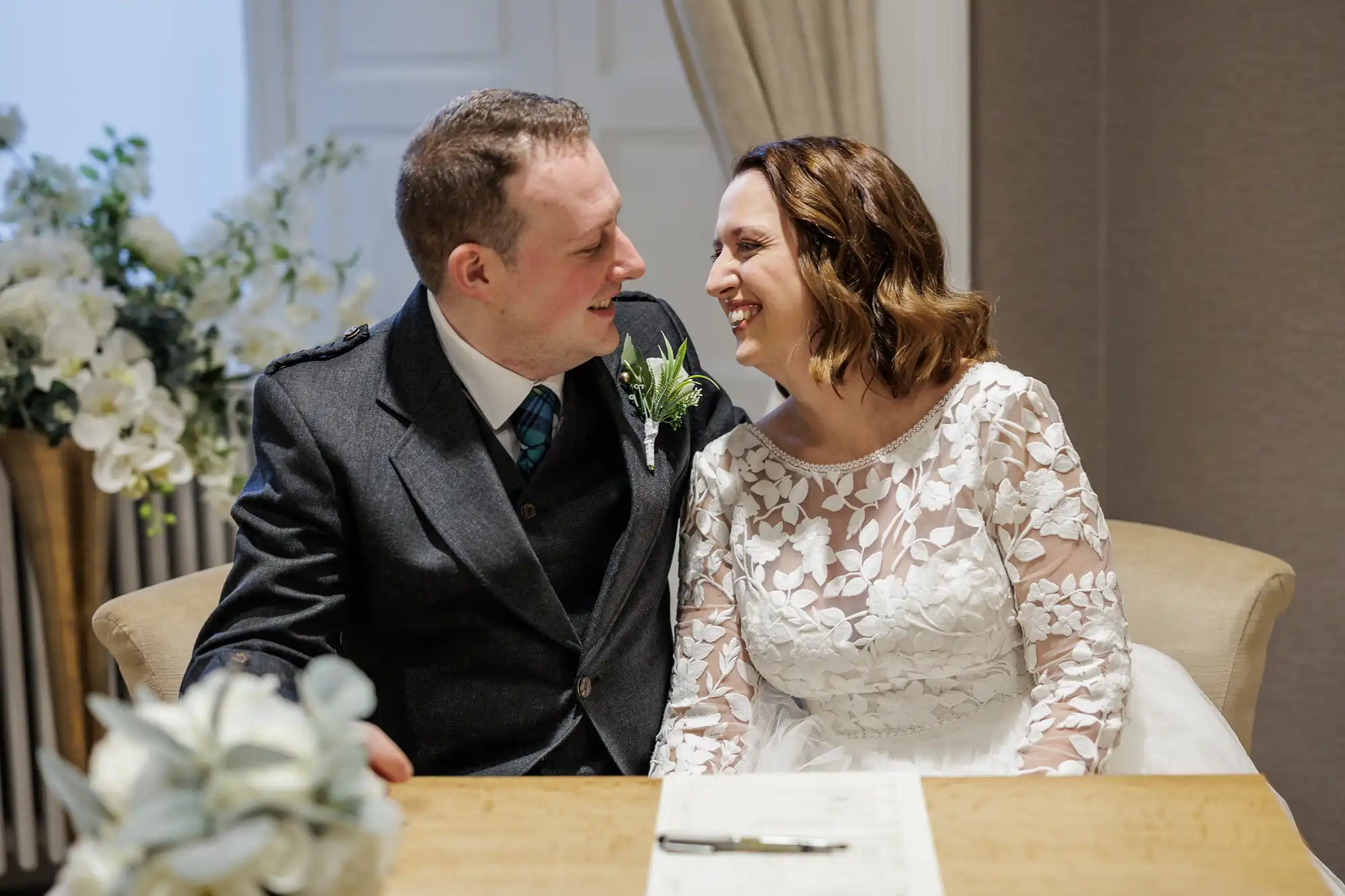 A smiling couple sit close together at a wooden table, dressed in wedding attire. The groom wears a dark suit with a tartan tie and buttonhole. The bride wears a white lace dress. A bouquet and a document with a pen are on the table; flowers decorate the background.