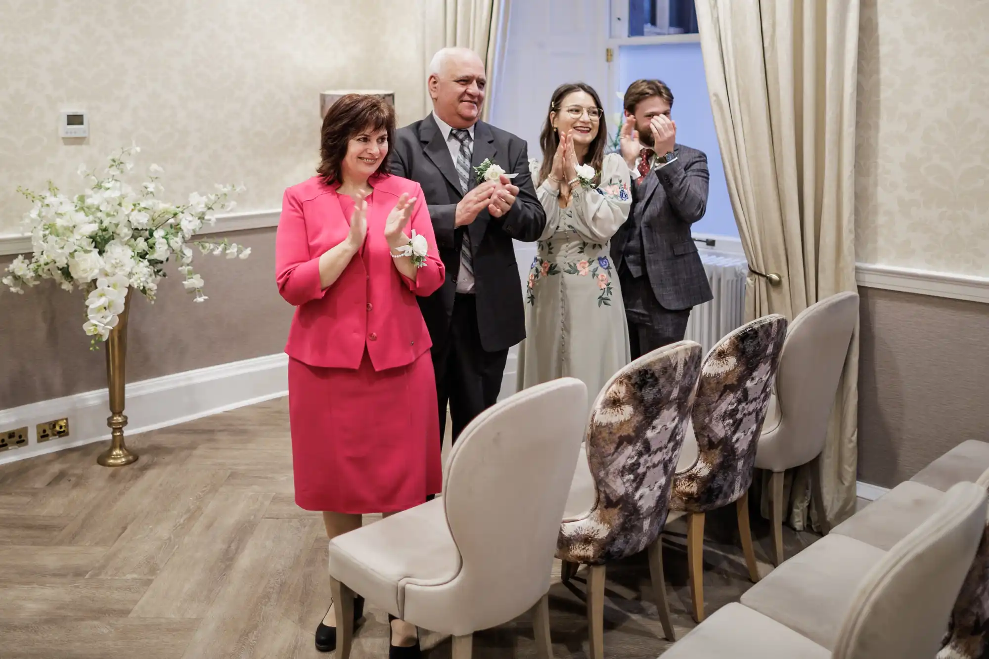 Four adults stand and clap in a small, elegant room with cream walls and floral decor. Three people smile, and one wipes tears from his eyes. Empty chairs face them, and sunlight filters through beige curtains. The mood appears joyful and emotional.