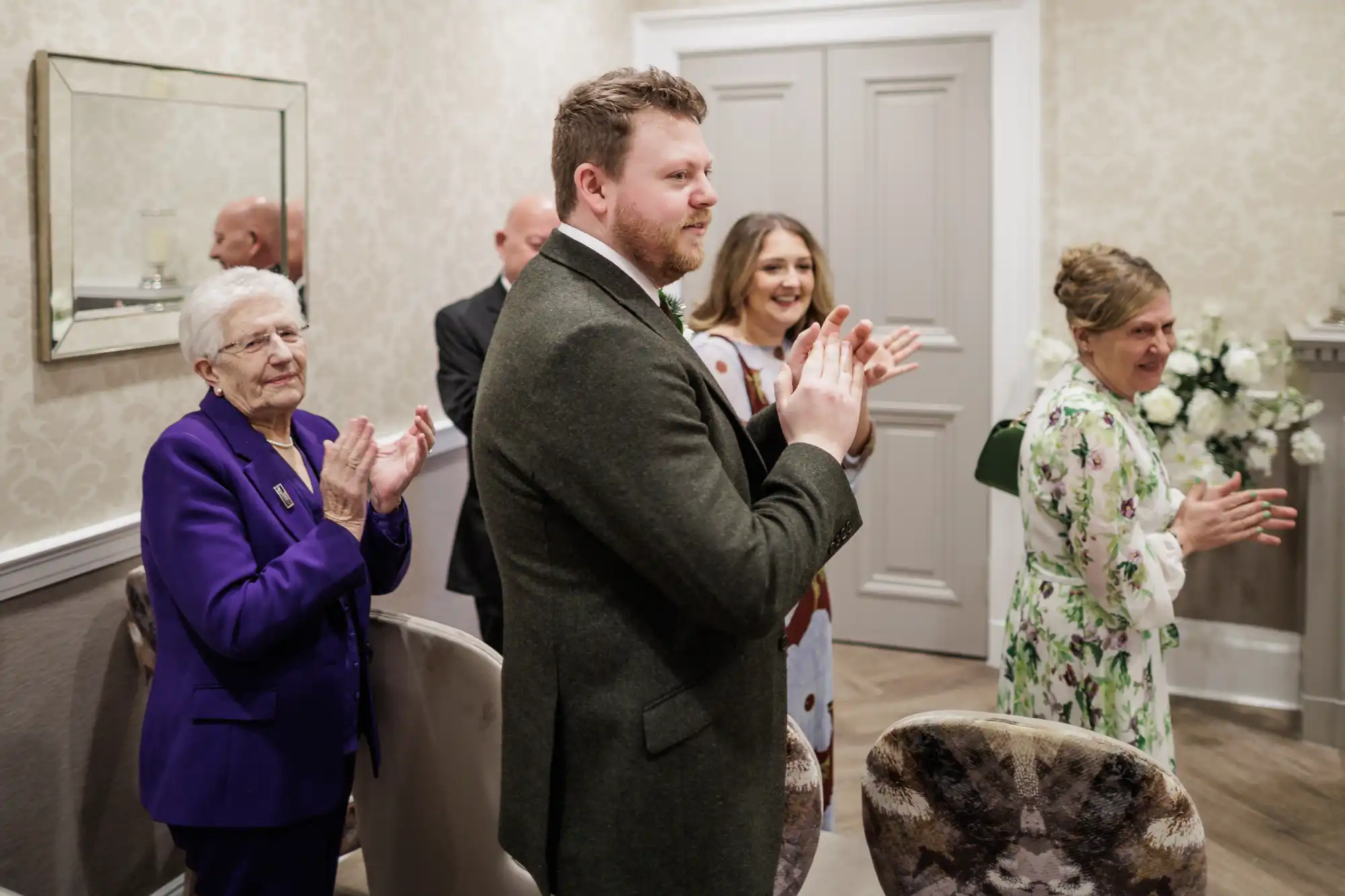 A group of people stands indoors, smiling and applauding. An elderly woman in a purple suit, a man in a green jacket, and two women in floral dresses are visible. The setting is elegant, with light-coloured walls, a mirror, and flower arrangements.