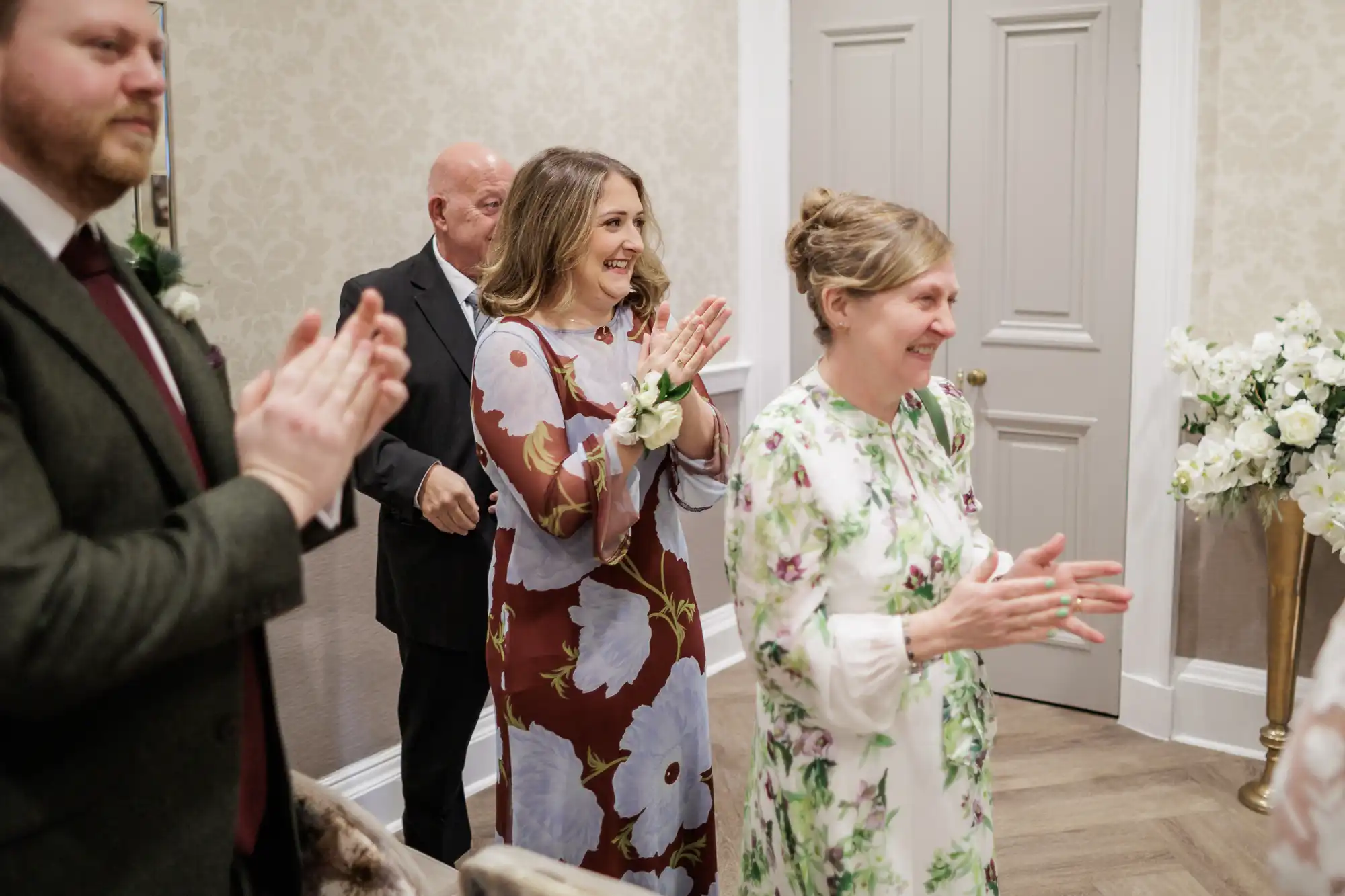 A group of people stand indoors clapping and smiling. Two women in floral dresses are at the front, with a man in a suit and an older man behind them. The room is decorated with light wallpaper, white doors, and white floral arrangements.