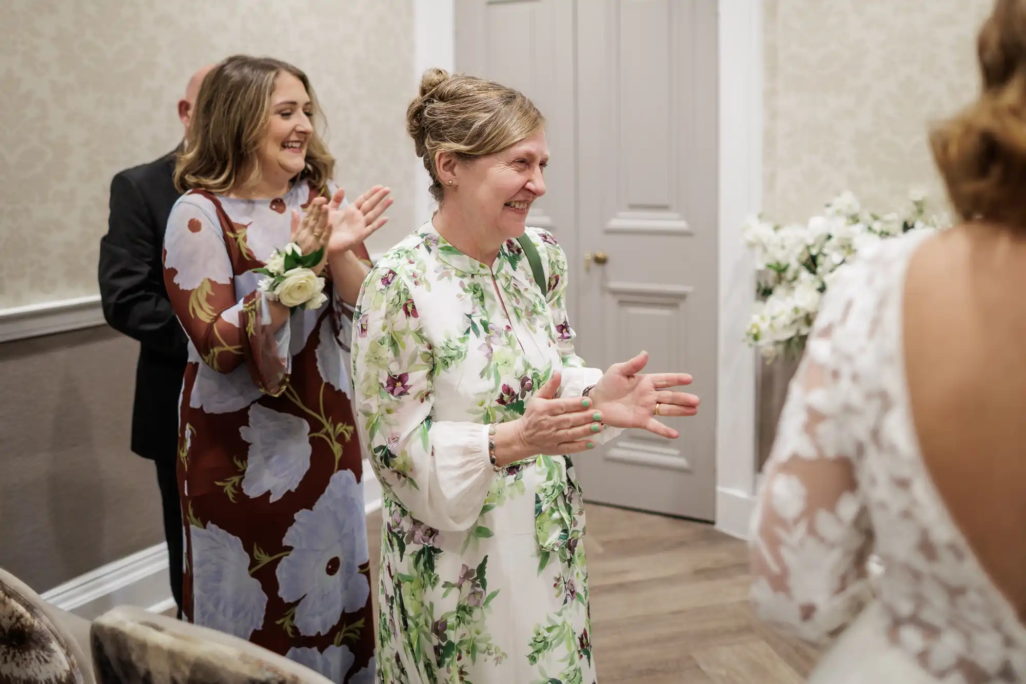 Two women in floral dresses smile and clap joyfully indoors, facing someone in a white lace dress, likely a bride. The setting appears elegant, with light wallpaper, a bouquet of white flowers, and soft lighting enhancing the celebratory mood.