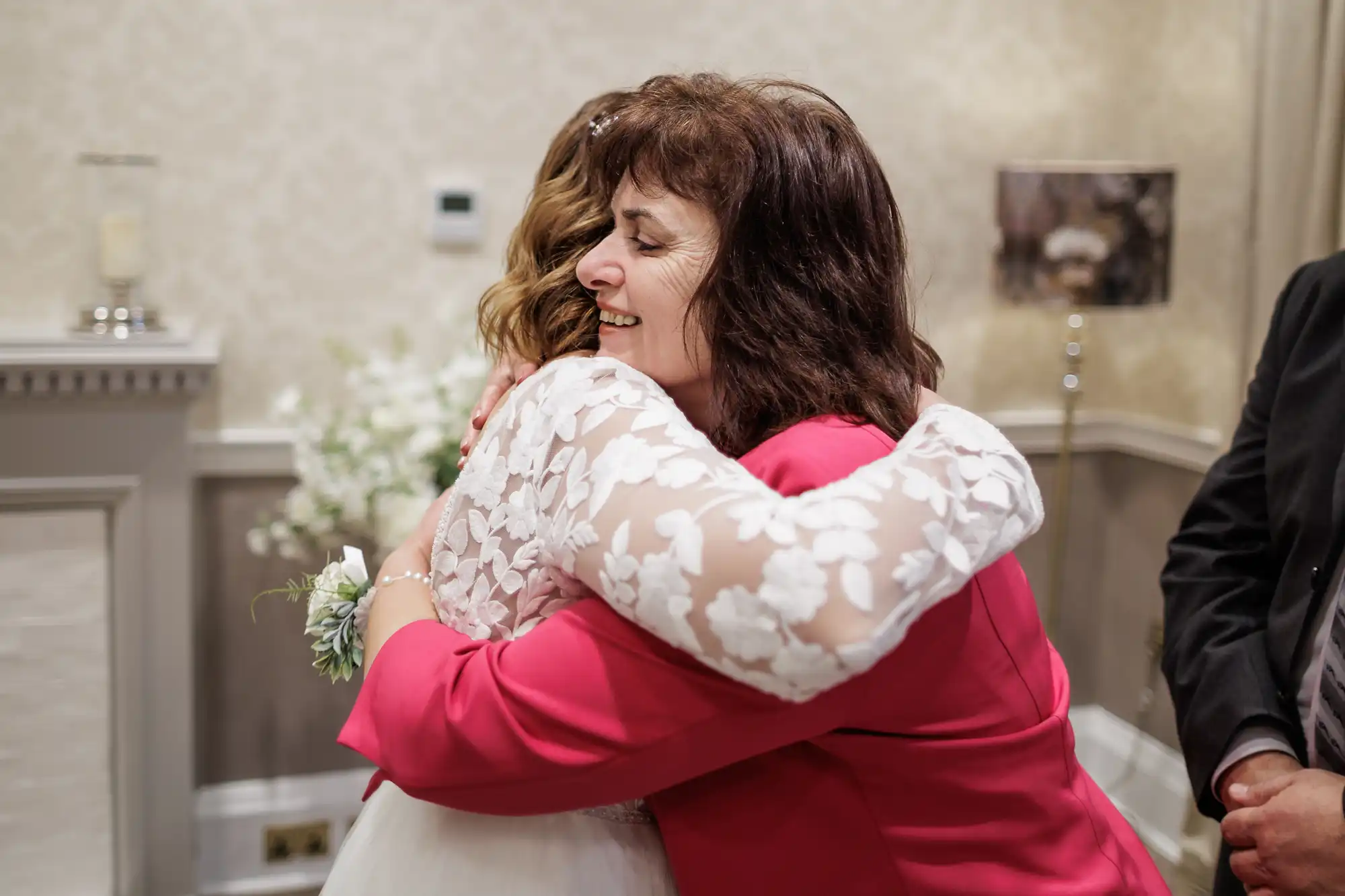 A woman in a pink jacket hugs a bride in a white lace dress. Both appear joyful, with the older woman's eyes closed and smiling. The setting is indoors with neutral décor, including a lamp, fireplace, and white flower arrangement in the background.