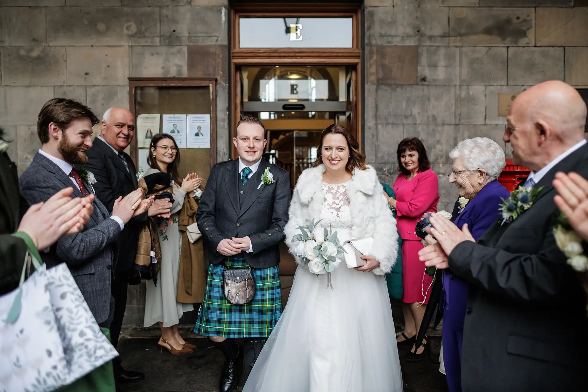 A bride in a white dress and fur shawl stands arm in arm with a groom in a grey jacket and green tartan kilt, both smiling as guests clap around them outside a stone building after their wedding ceremony.
