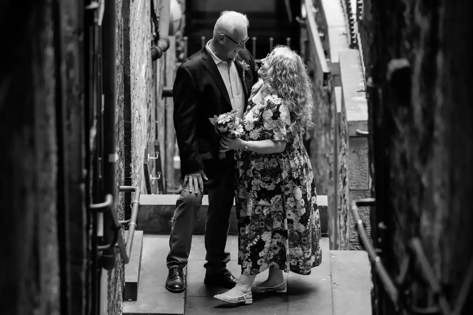 A couple stands close together in a narrow alleyway, gazing into each other’s eyes. The woman holds a bouquet and wears a floral dress, while the man wears a suit jacket and glasses. The scene is in black and white.