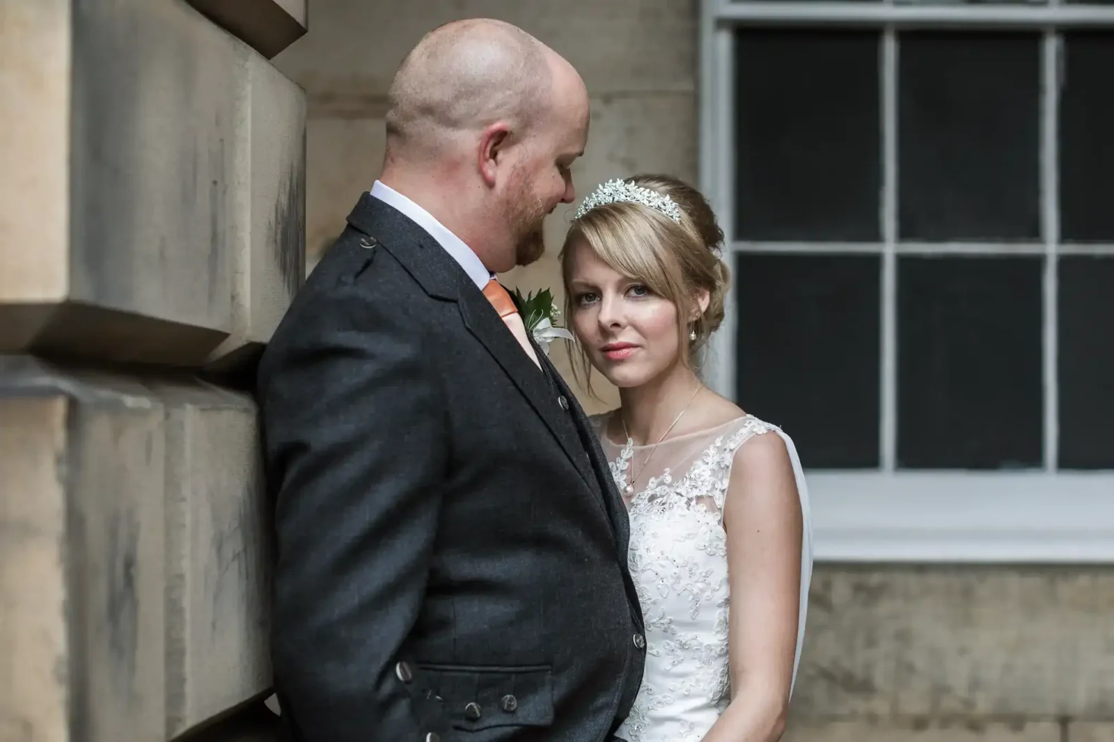 A bride and groom stand close together against a stone wall, with the groom looking at the bride and the bride facing the camera.