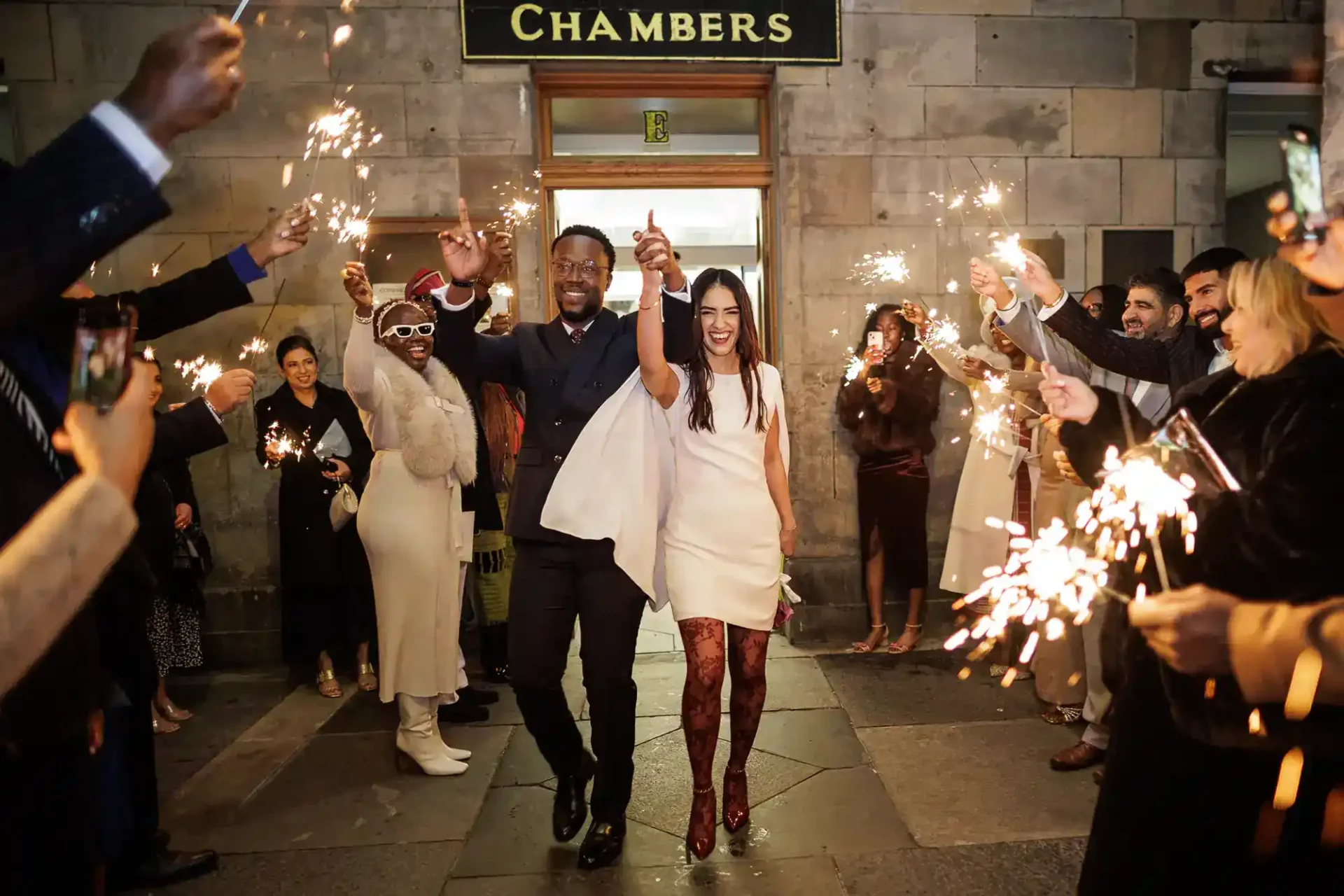 A couple walks hand in hand through a crowd holding sparklers, celebrating outside a building with a "Chambers" sign above the door at City Chambers Edinburgh.