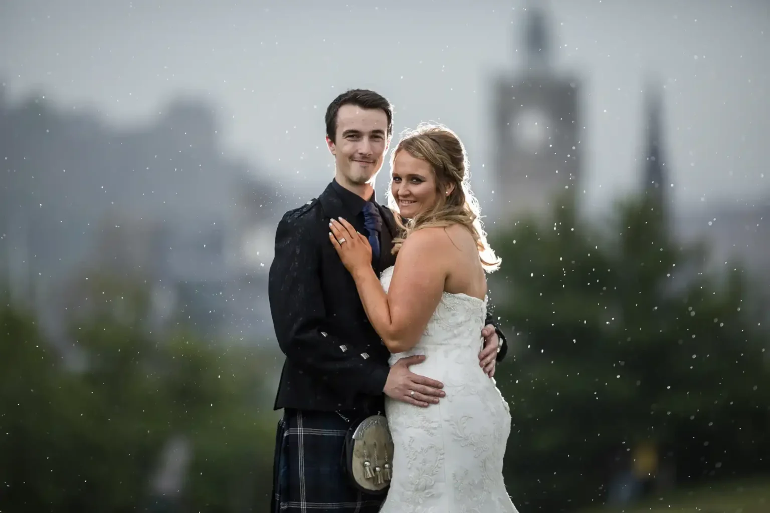 A bride and groom stand close together outdoors in the rain, smiling at the camera, with blurred greenery and buildings in the background.