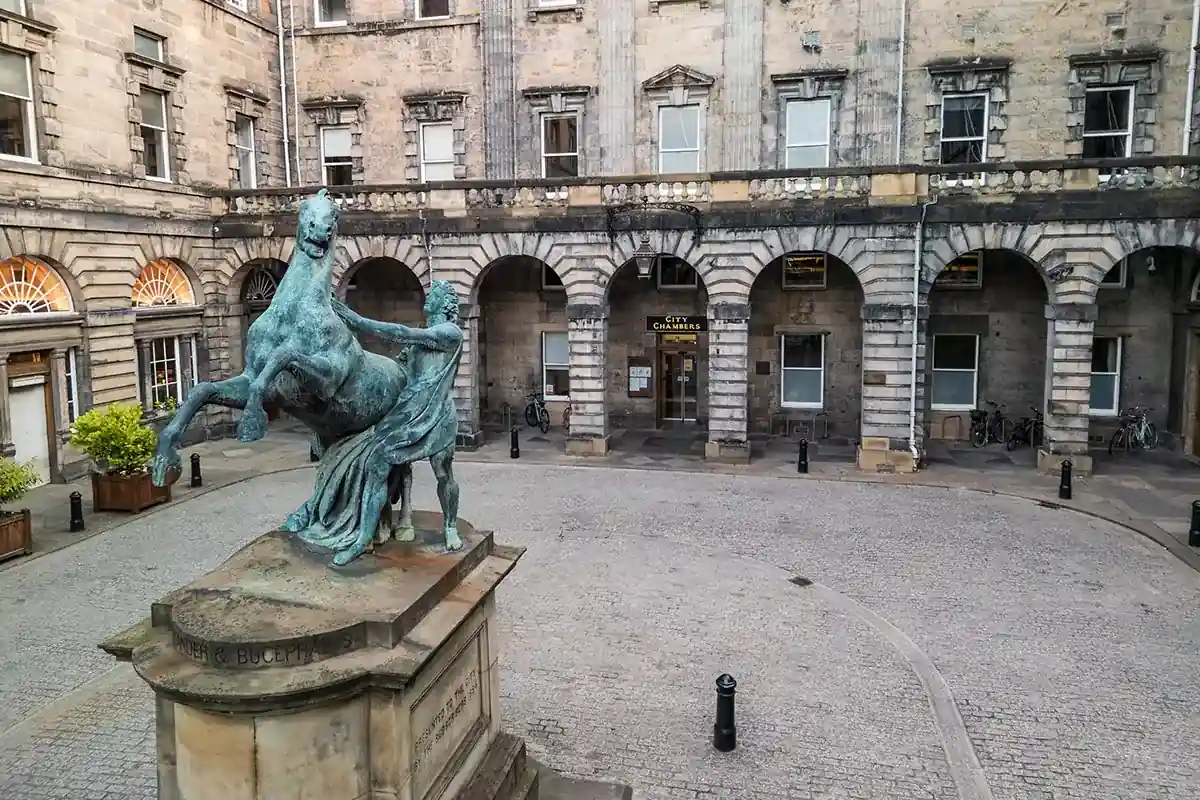 A weathered bronze statue of a horse and rider stands on a stone plinth in a quiet, cobbled courtyard, bordered by an old, grey stone building with arches, columns, and rectangular windows. A small tree in a planter sits near one wall.