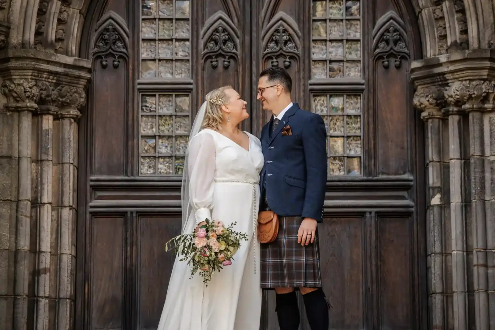 A bride in a long white dress and veil holds a bouquet and smiles at a groom in a blue jacket, checked kilt, and sporran. They stand close together, gazing at each other, in front of an ornate, dark wooden door with gothic arches and patterned glass.