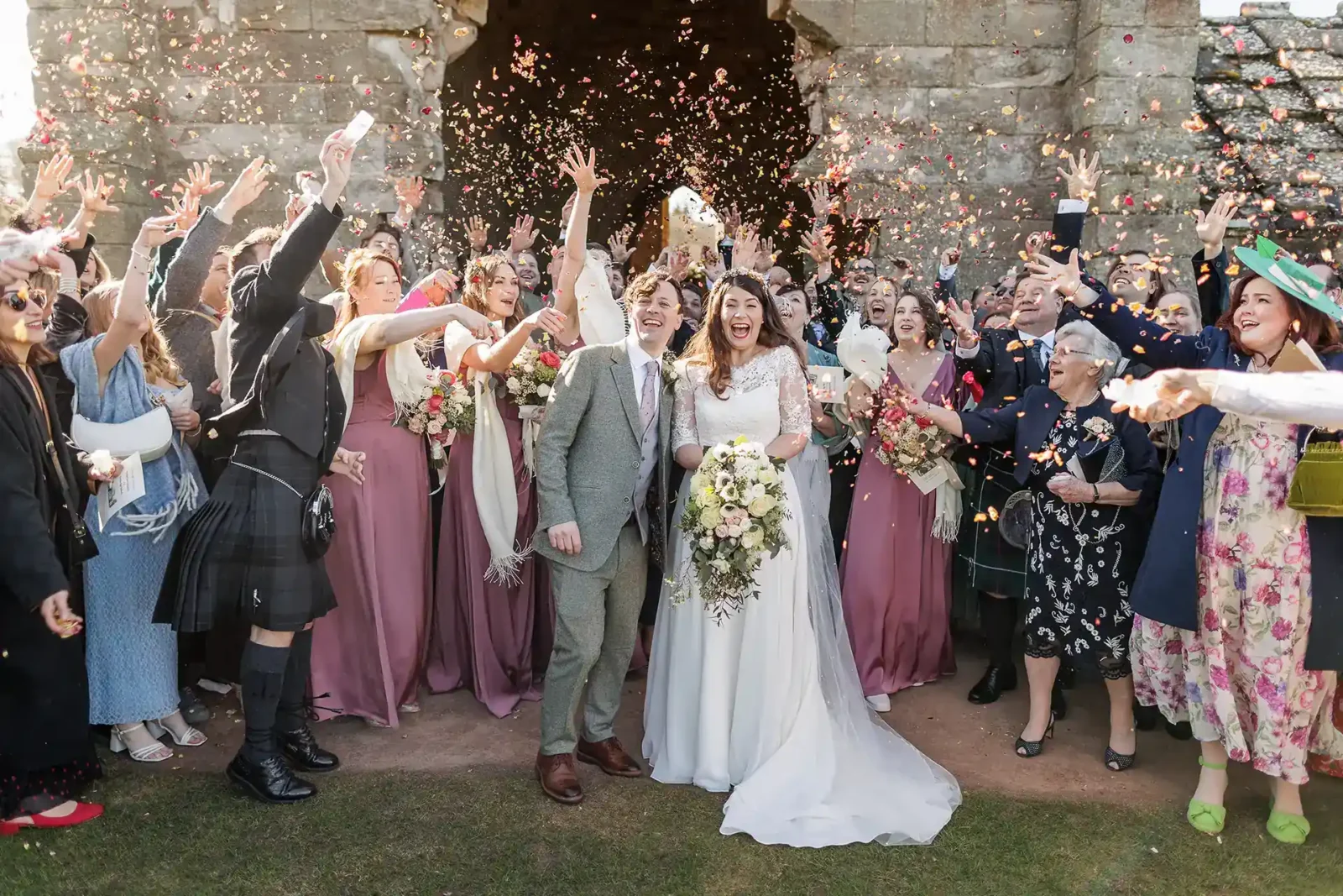 Award-winning Edinburgh wedding photographer team Jonathon and Claire - Jonathon Fowler Photography. A bride and groom stand smiling surrounded by guests throwing confetti outside a stone building at a wedding celebration.