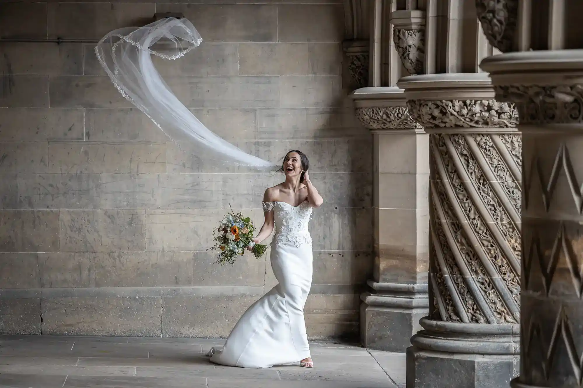 Edinburgh wedding photographer photography session of a bride in a white dress stands indoors, holding a bouquet, as her veil floats in the air behind her near ornate stone columns at Fettes College, Edinburgh.