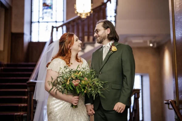European Room winter elopement photo of a bride in a beaded white dress and veil holds a bouquet, smiling at the groom in a green suit with a buttonhole. They stand hand in hand indoors, in front of a wooden staircase and stained glass window, looking happily at each other.