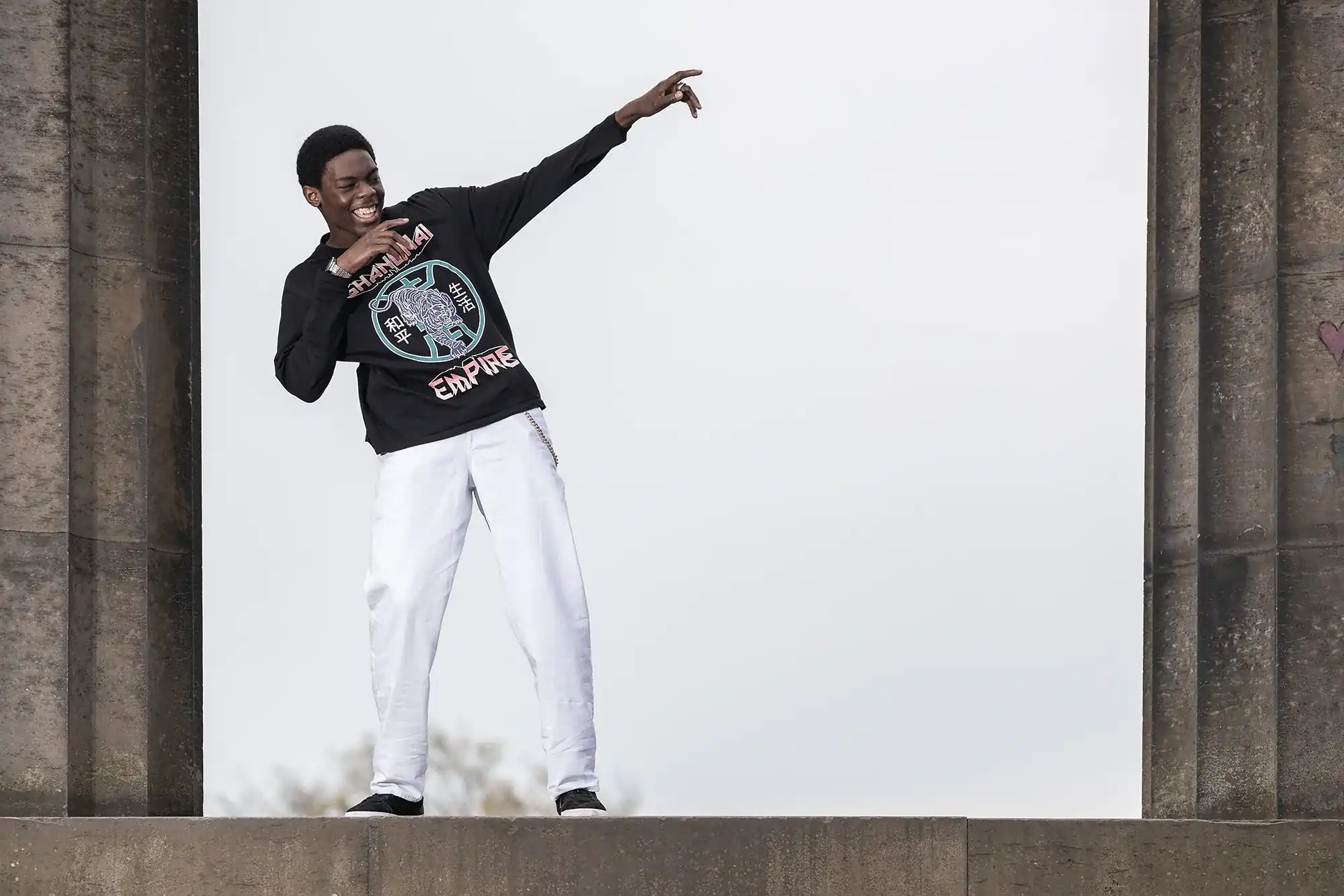 A person wearing a black long-sleeve shirt and white pants poses with a smile, pointing upwards with their right hand. They are standing between large stone structures against a cloudy sky.