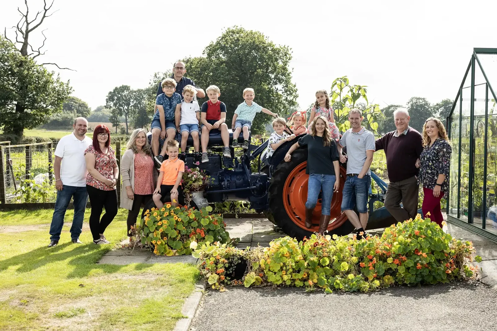 A group of people, including adults and children, stand and sit around a blue tractor surrounded by foliage in an outdoor garden setting.