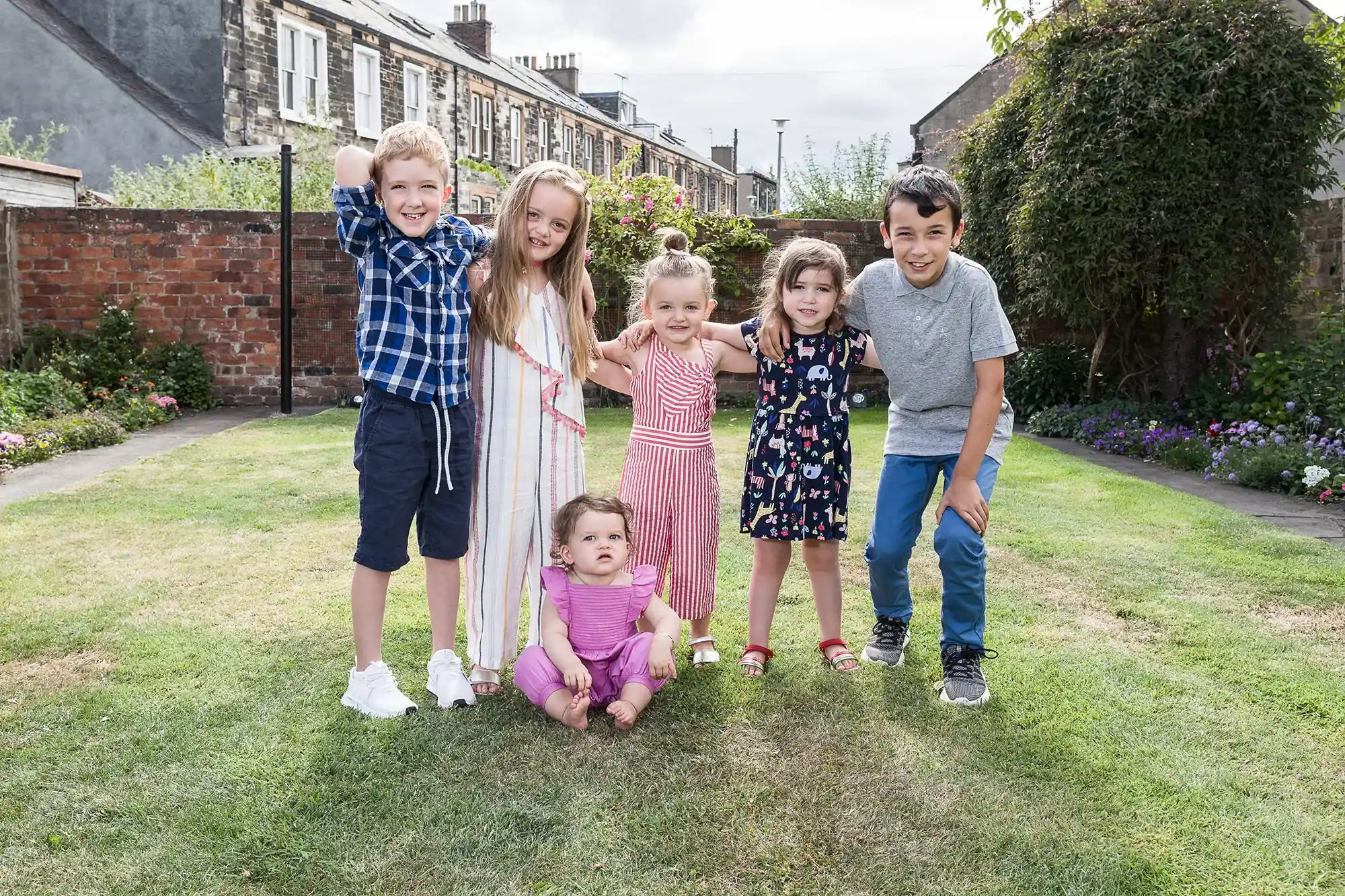 Six children stand and sit on grass in a backyard, posing for a photo. There are four girls and two boys. Stone houses and a brick wall are visible in the background.