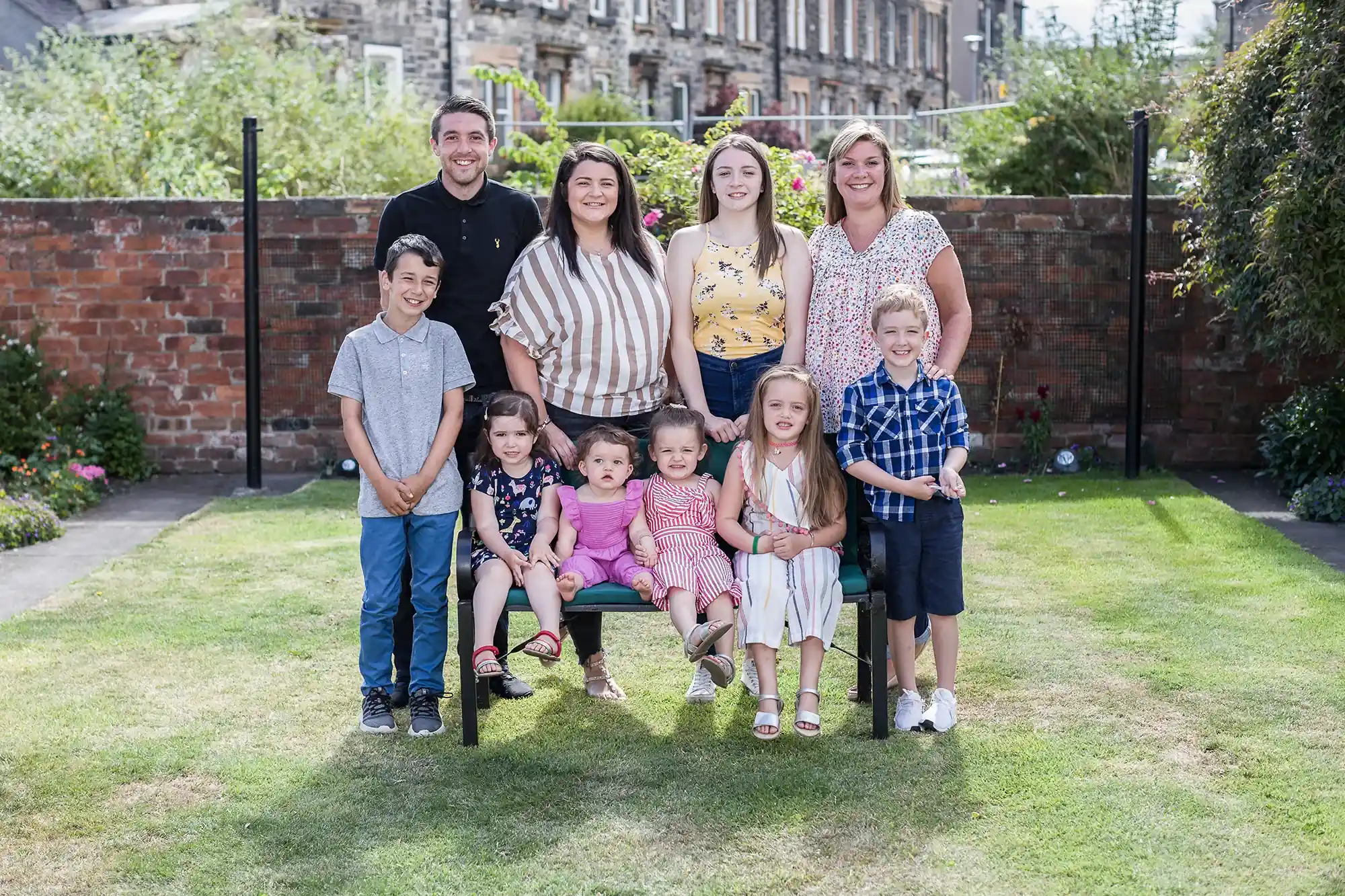 A group of four adults and six children pose for a photo in a garden. Five children are seated on a bench, and the adults and one child stand behind them. A brick wall and buildings are in the background.