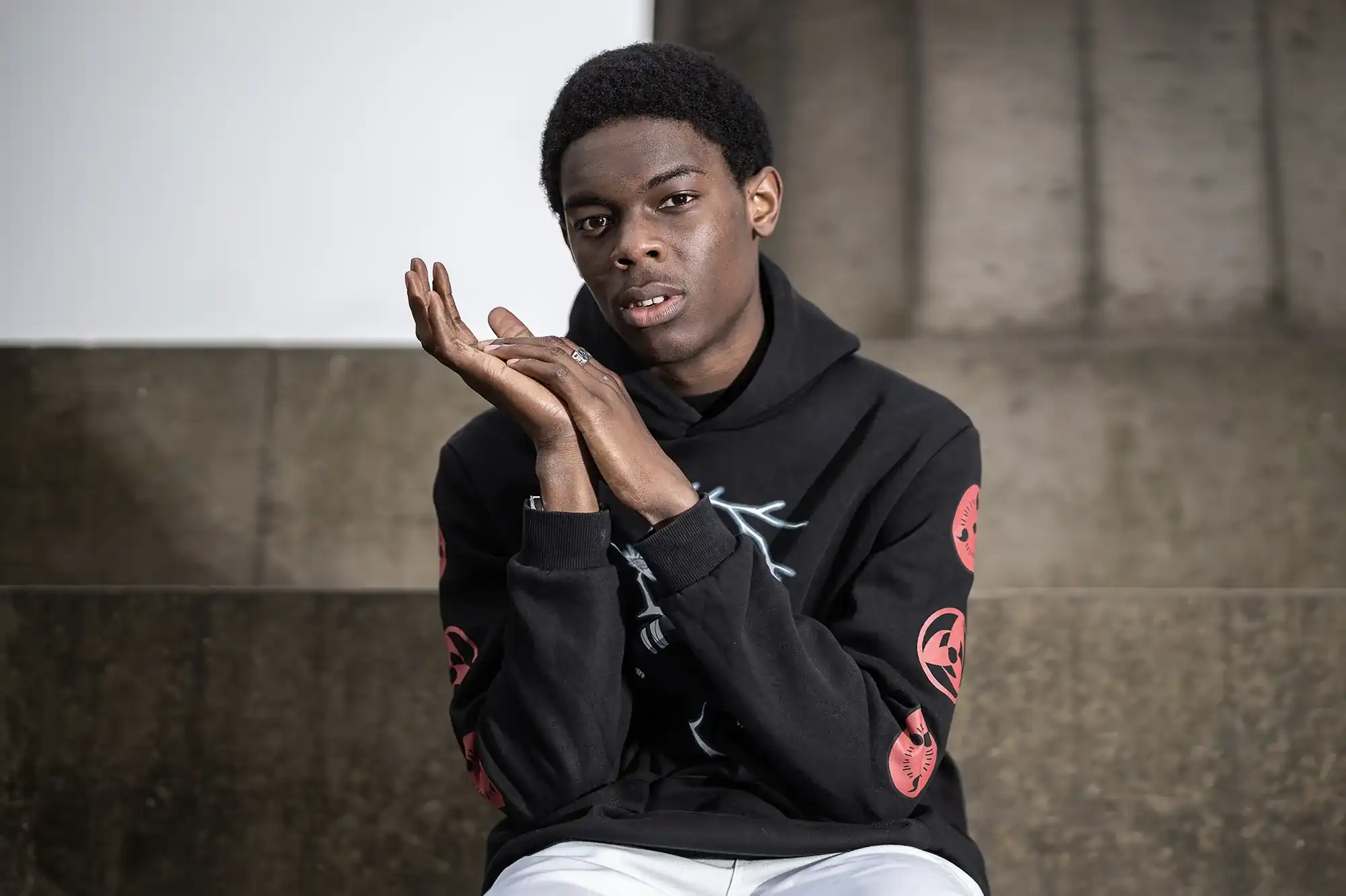 A person wearing a black hoodie with red designs sits in front of a concrete wall, clasping their hands together and gazing into the camera.