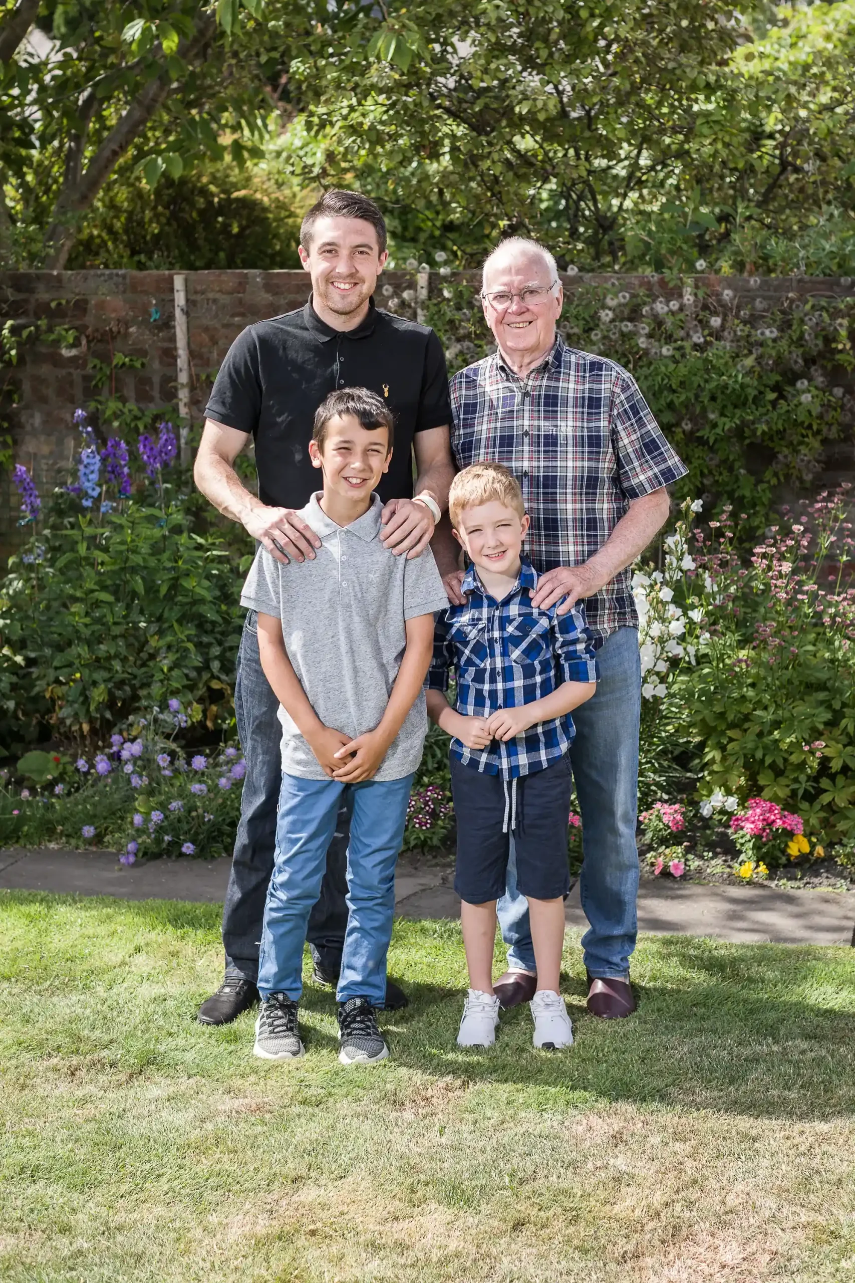 A group of four people, consisting of two adults and two children, standing together outdoors in a garden with flowers and greenery in the background.