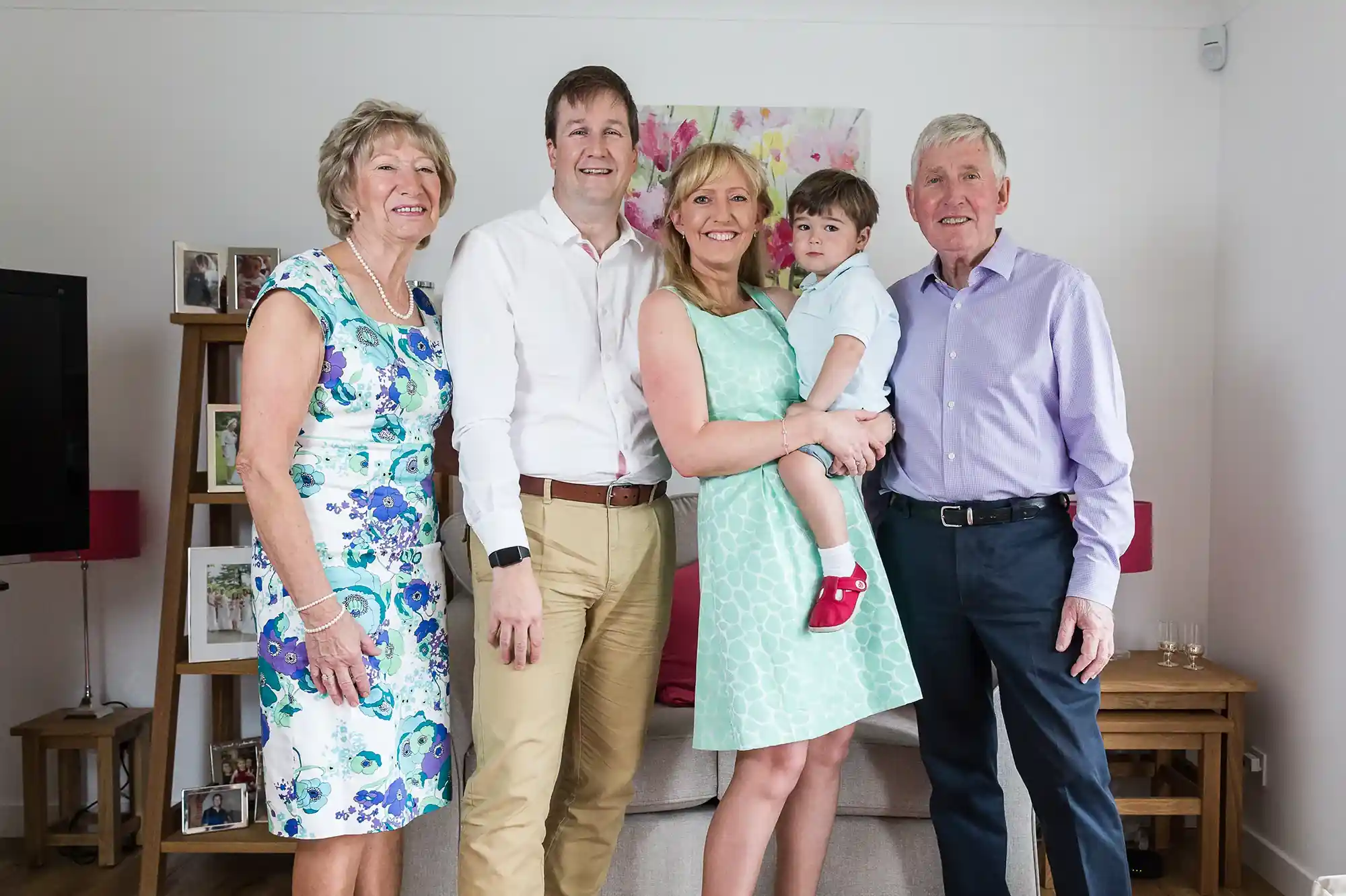 A group of five people, including a child, standing indoors and smiling. They are dressed in semi-formal attire and appear to be in a living room with framed photos and furniture in the background.