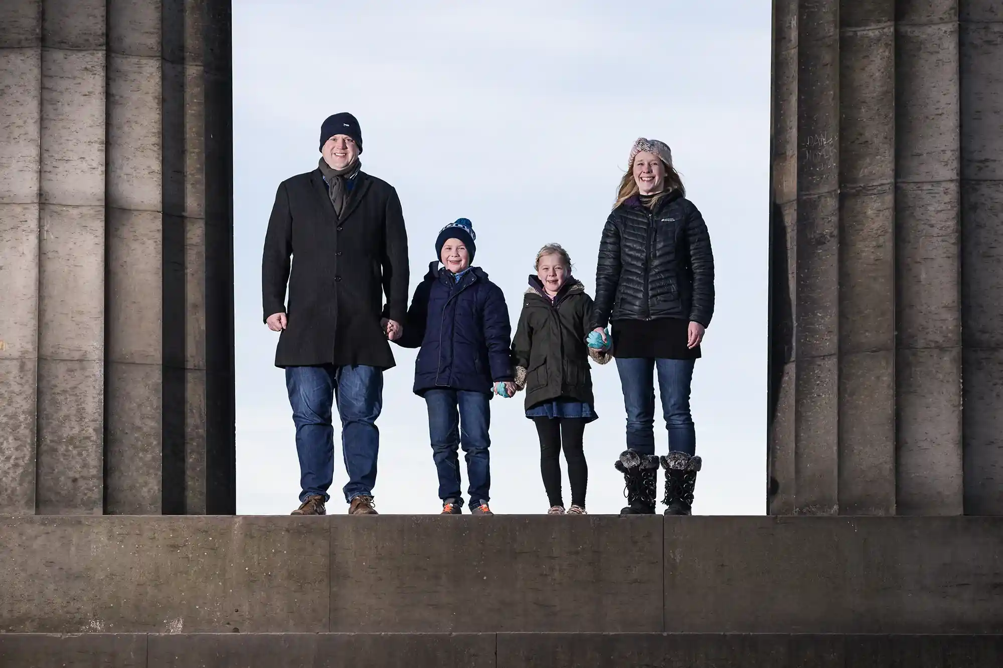 Four, dressed in winter clothes, stand between large stone pillars. The parents stand on the sides, holding hands with their two children in the middle.