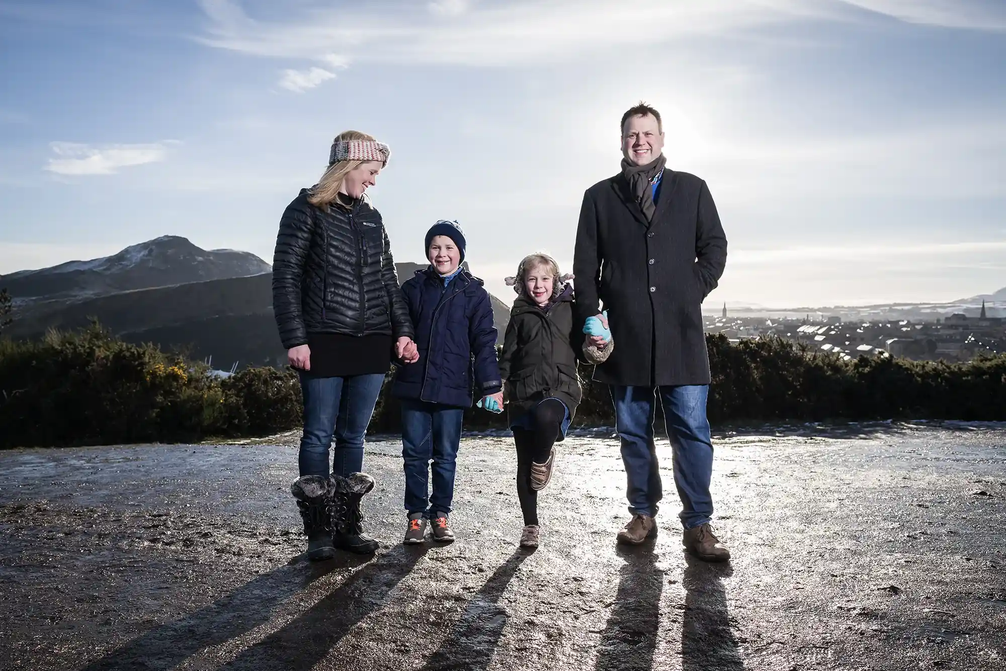 two adults and two children, stand outdoors on a paved path with a scenic mountainous backdrop. The adults wear coats, and the children wear jackets and hats. One child lifts a leg playfully.