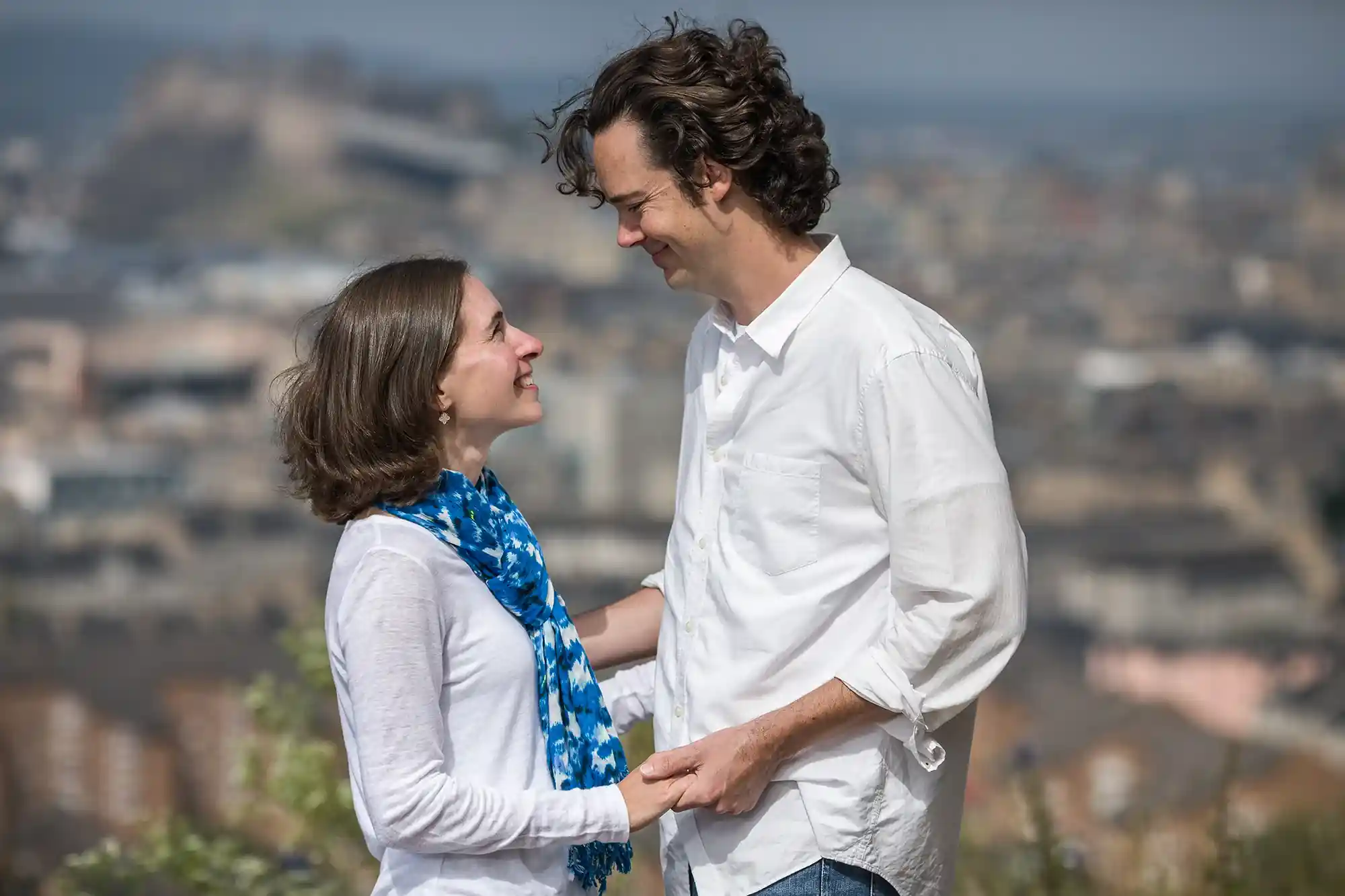 A man and woman, both wearing white shirts, stand facing each other and smiling while holding hands. They are outside with a blurred cityscape in the background.