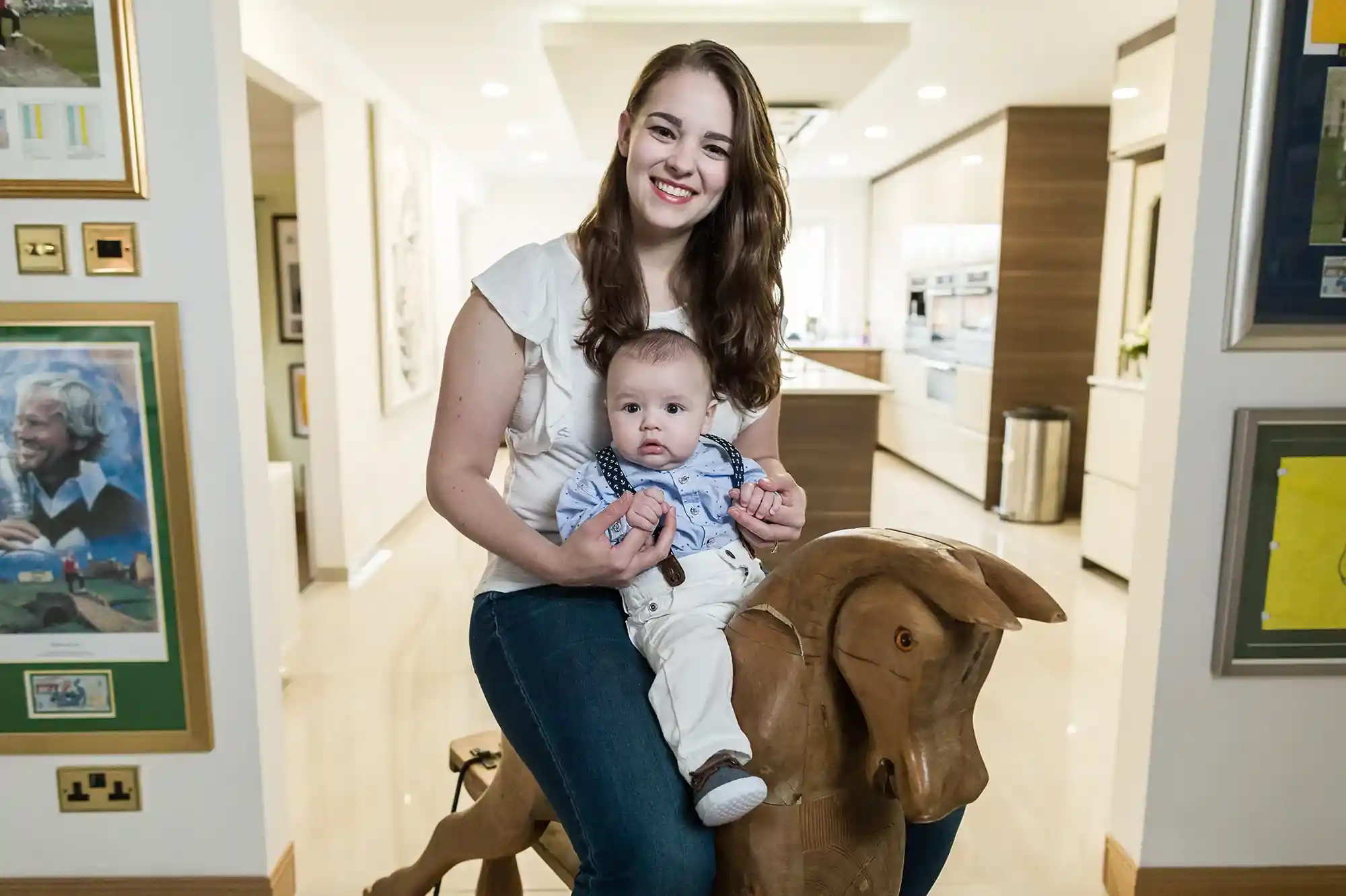 A woman smiles while holding a baby seated on a wooden rocking horse in a well-lit room with framed pictures on the walls.