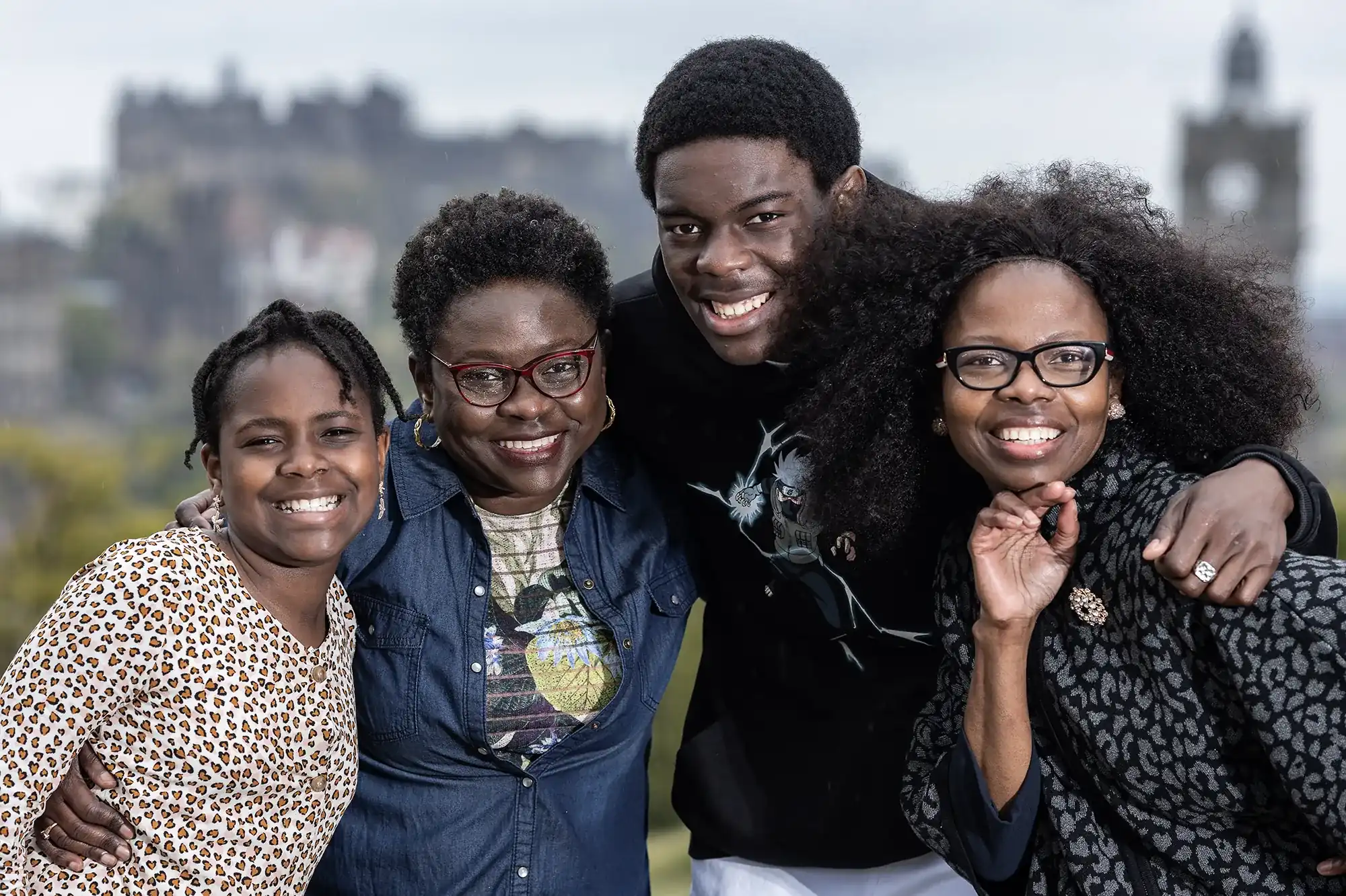 A group of four people stand close together outdoors, smiling at the camera with a castle visible in the background.