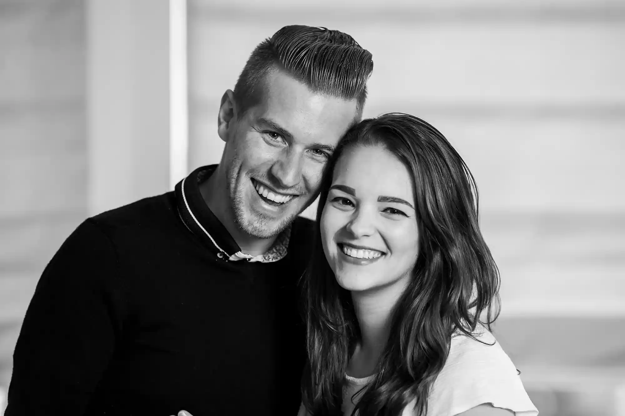 A monochrome photo of a smiling man and woman standing closely together, both looking directly into the camera.