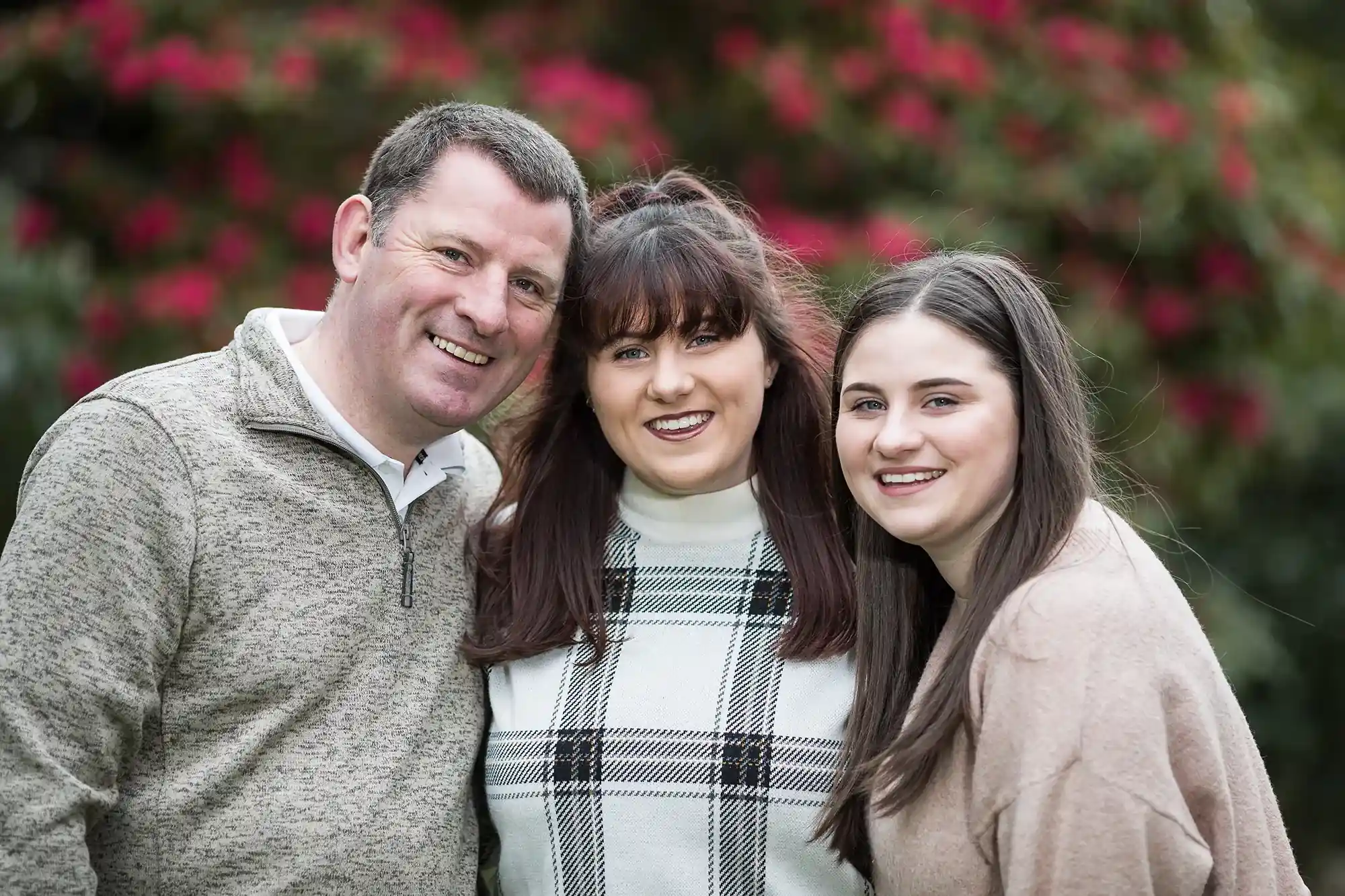 Three people posing together outdoors in front of greenery and red flowers, all smiling at the camera.